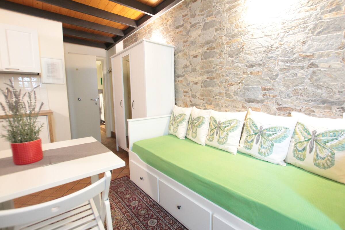 A bedroom with a stone wall, a bed with butterfly pillows, a white wardrobe, and a small table with a red pot.