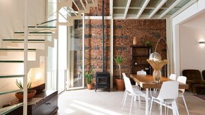Una sala de estar moderna con una escalera de cristal, una pared de ladrillo, una chimenea y un comedor con sillas blancas.