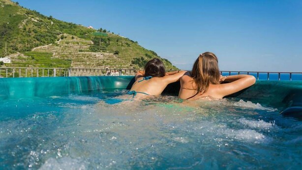 Two people relaxing in a hot tub with a view of a green hillside and a clear blue sky. - Palazzo Cocò (Maiori)