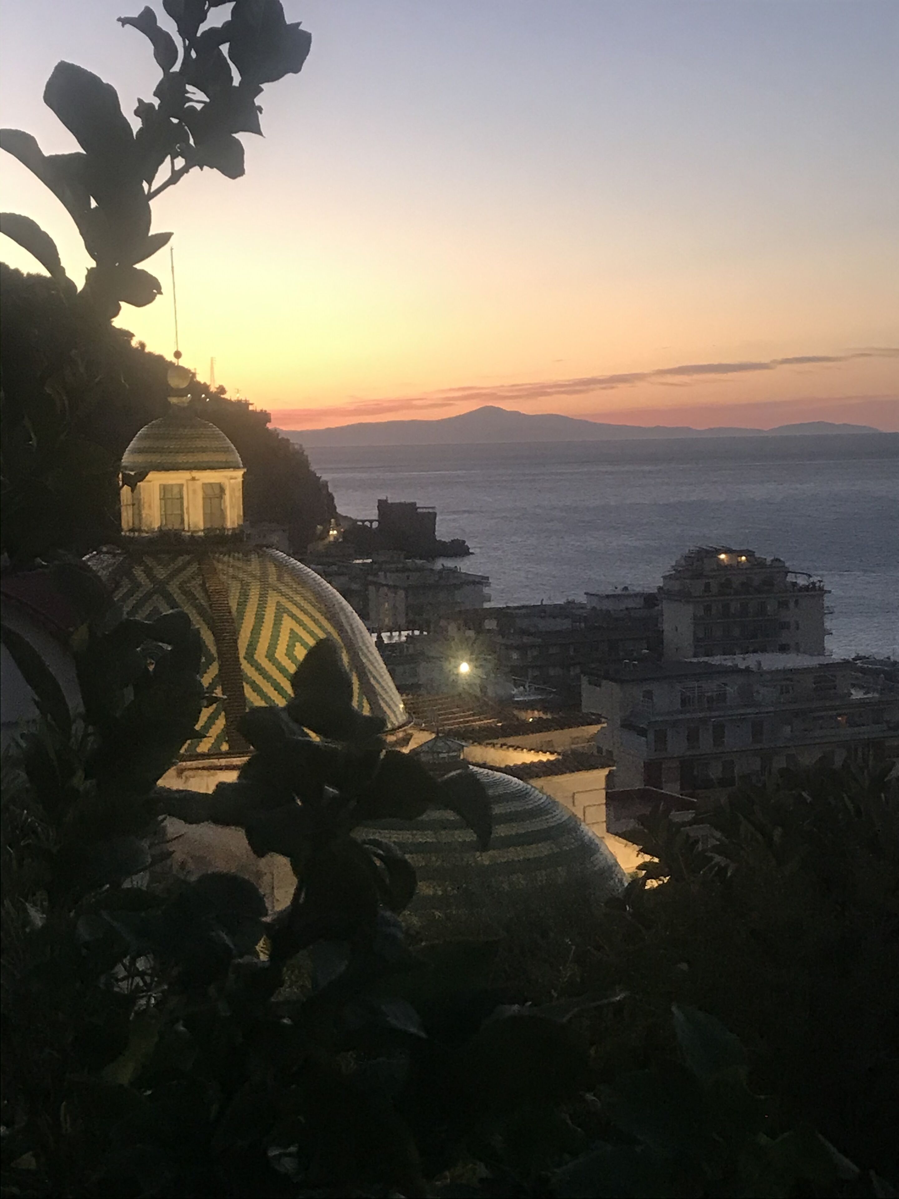 A coastal cityscape at sunset with prominent domed buildings and a view of the sea.