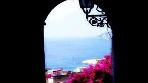 A view through an arched window overlooking a coastal area with buildings and pink flowers. - Palazzo Cocò (Maiori)