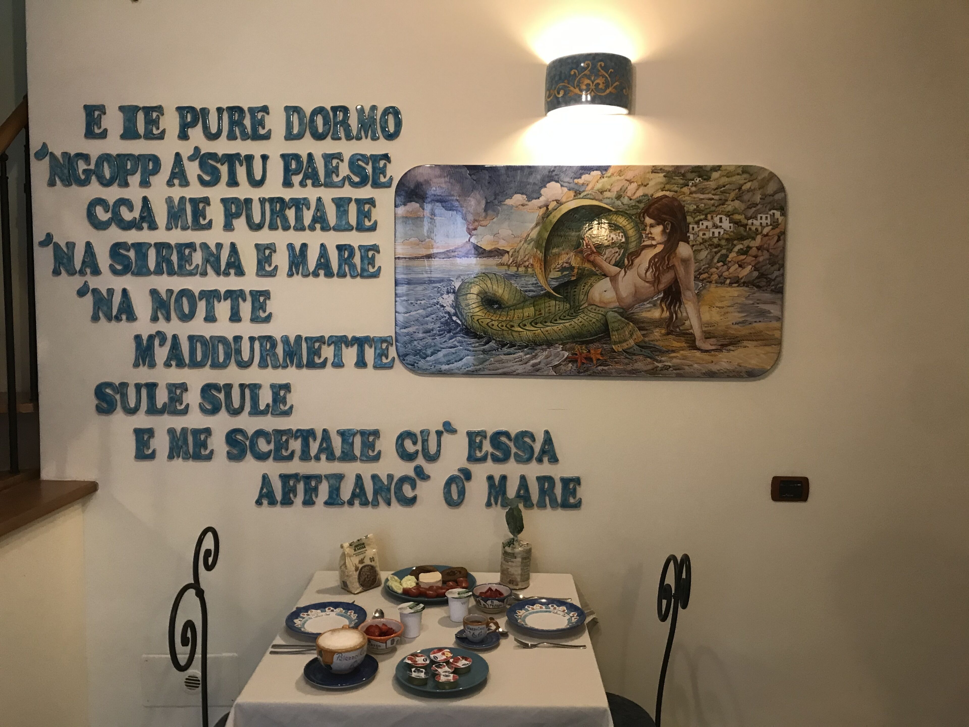 A wall with a poem in Italian, a painting of a mermaid, and a table with breakfast items.