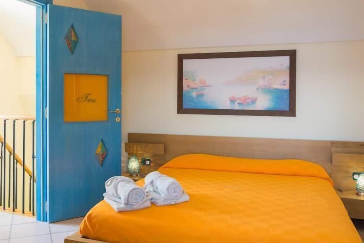 A hotel room with a bed, a blue door with a 'Tea' sign, a painting of boats, and a staircase.