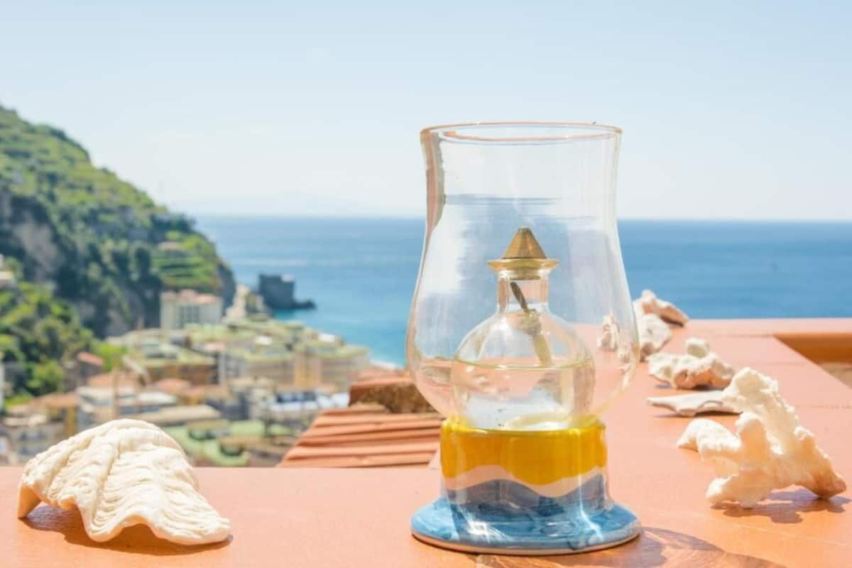 A clear glass bottle with a yellow liquid, placed on a wooden table with seashells, overlooking a coastal town and the sea.