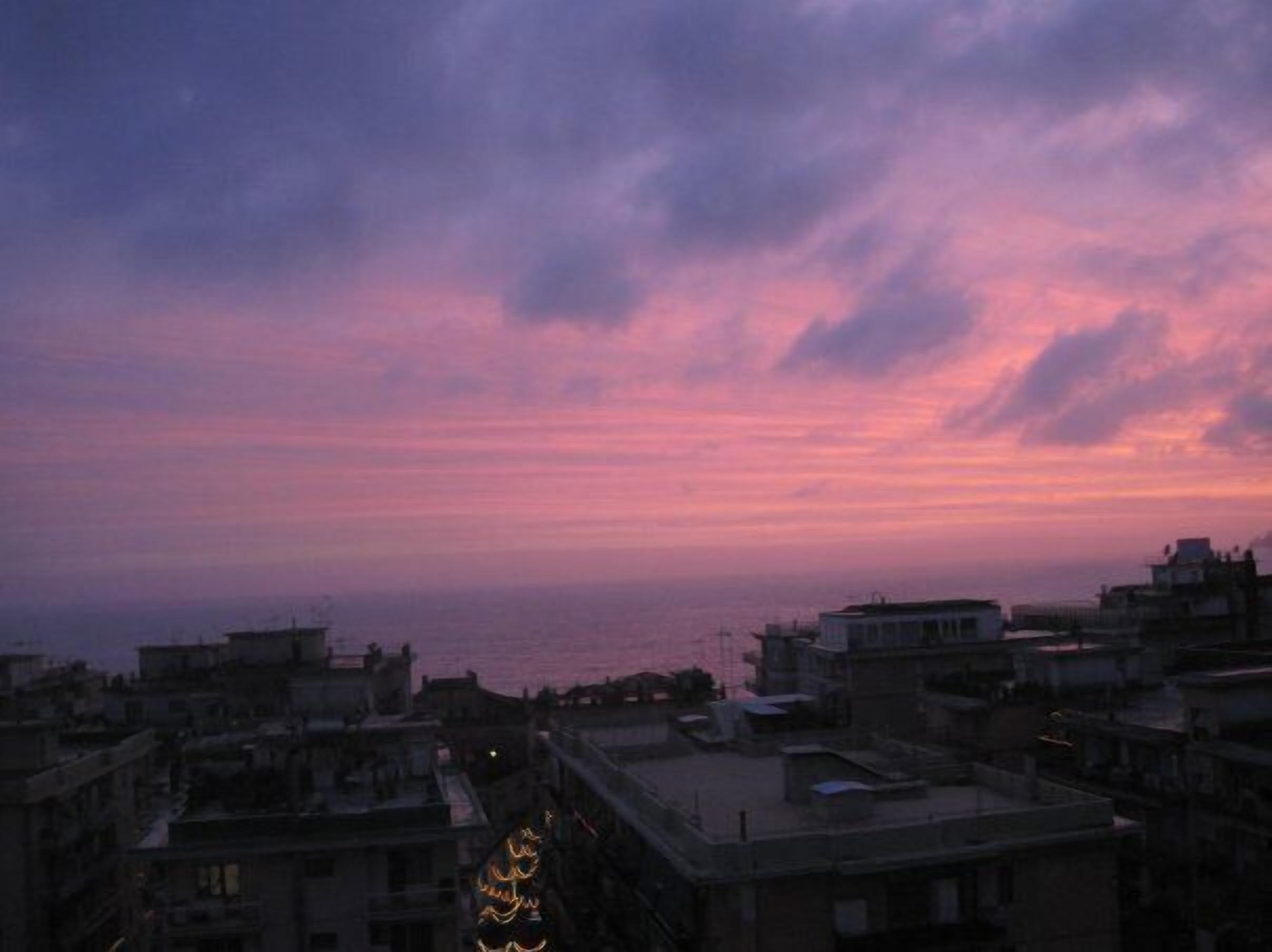 A cityscape at dusk with buildings and a view of the sea.