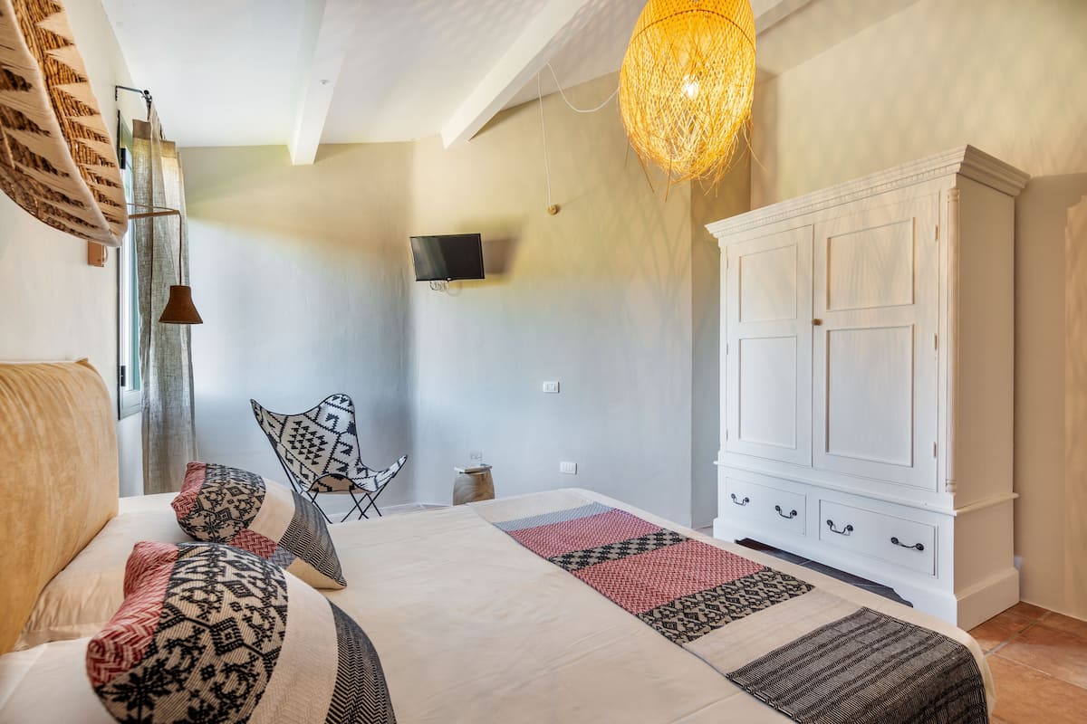 Un dormitorio con una cama, almohadas estampadas, un armario blanco, un televisor y una lámpara colgante.