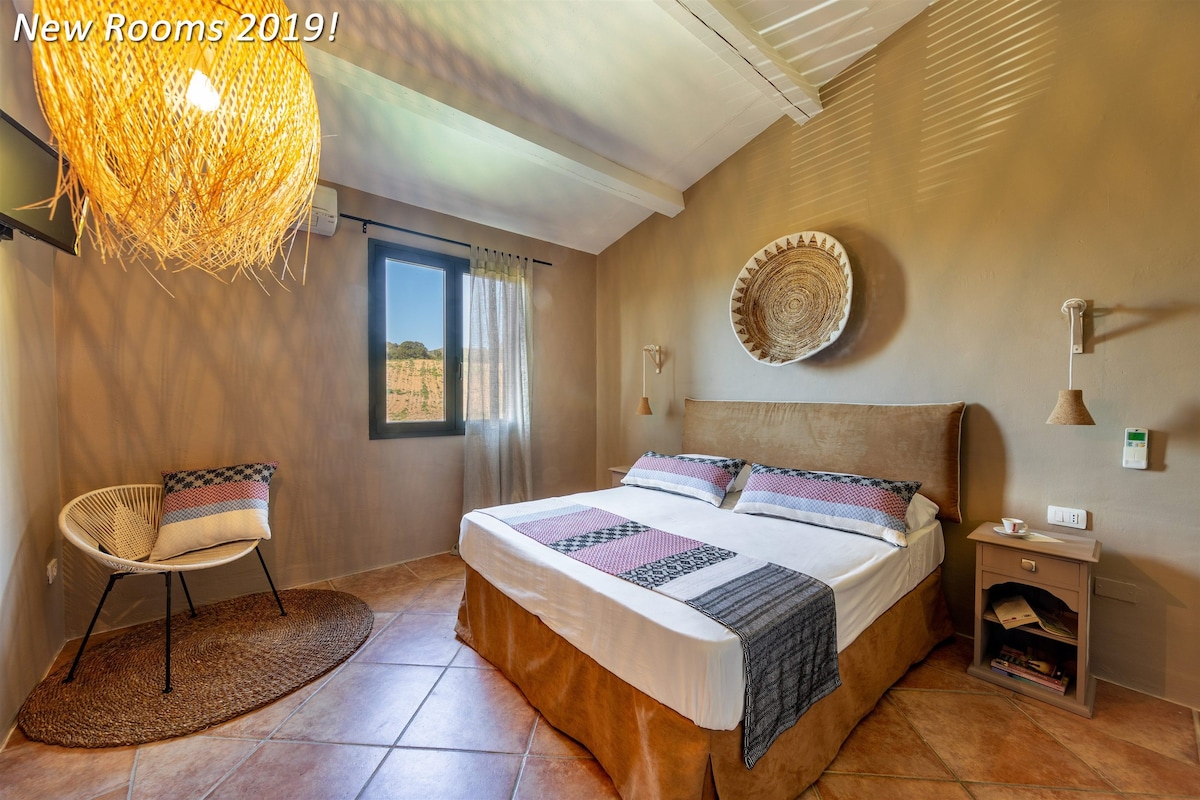 A bedroom with a large bed, a woven chair, a woven rug, a wall-mounted lamp, and a view of the outdoors.