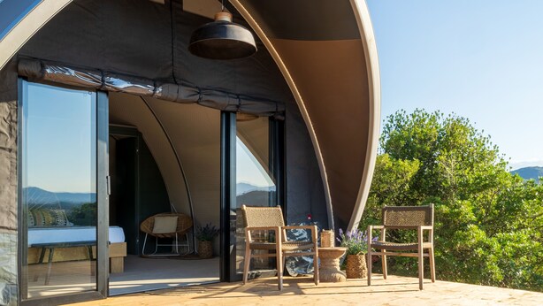 A modern, curved-roofed structure with a glass door leading to an interior space. Inside, there is a bed, a wicker chair, and a table with a vase of flowers. - Hotel Glamping Borgo di Campagna (Loiri Porto San Paolo)
