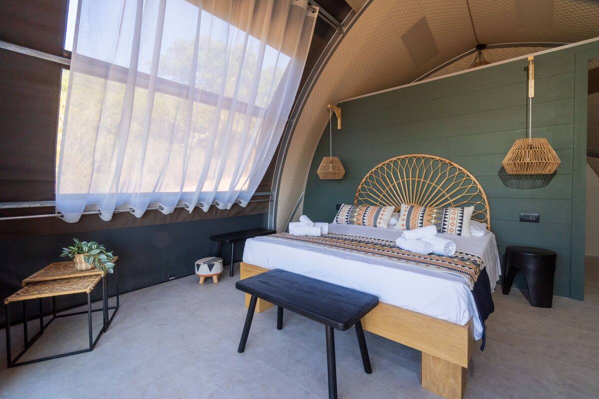 Un dormitorio moderno con una cama grande, un banco, una mesita y una lámpara colgante.