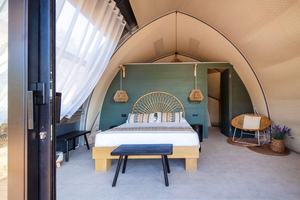 Una habitación con carpa, una cama, un banco, lámparas colgantes y una silla de mimbre.
