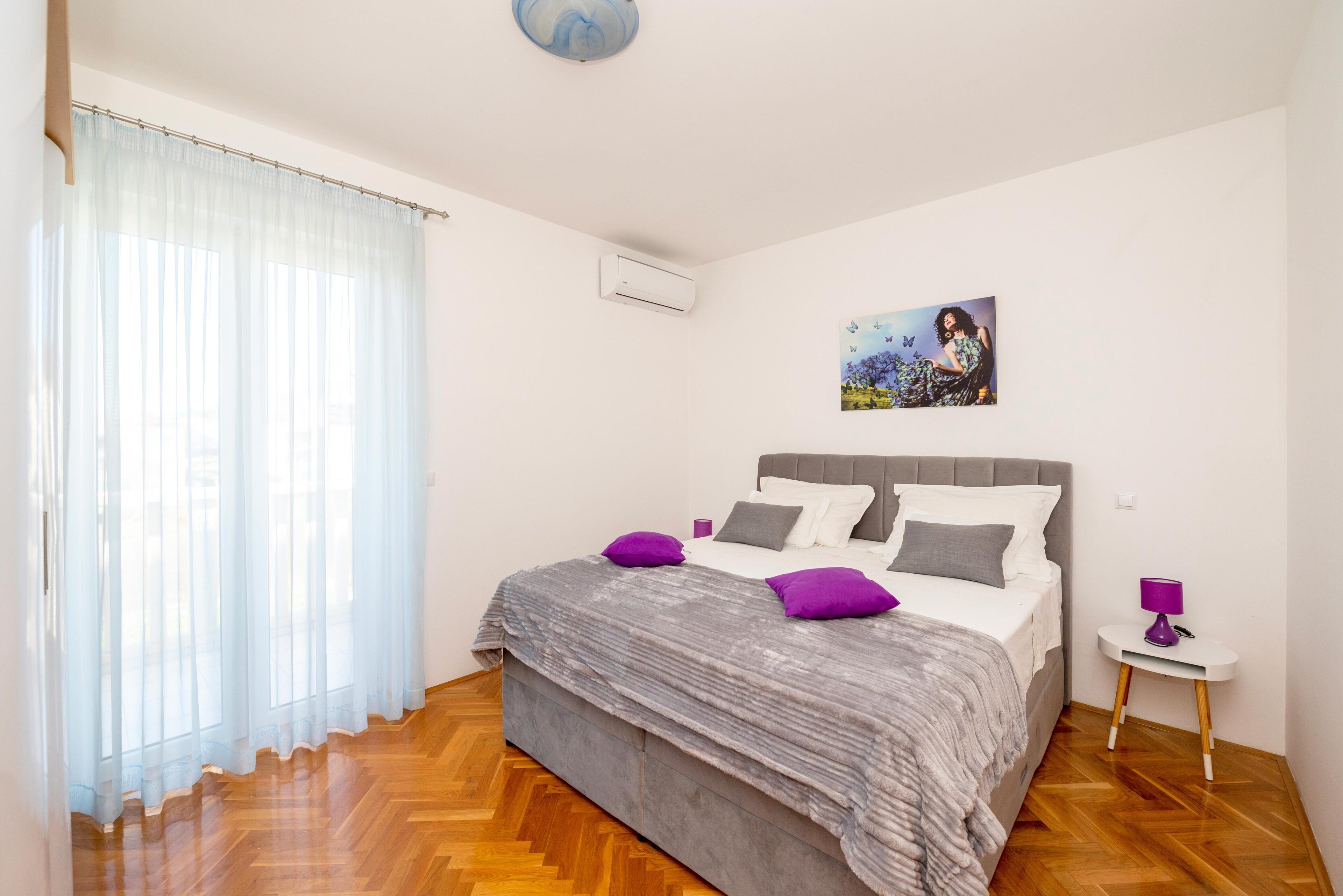 A bedroom with a bed, a nightstand, a painting, and a window with sheer curtains.