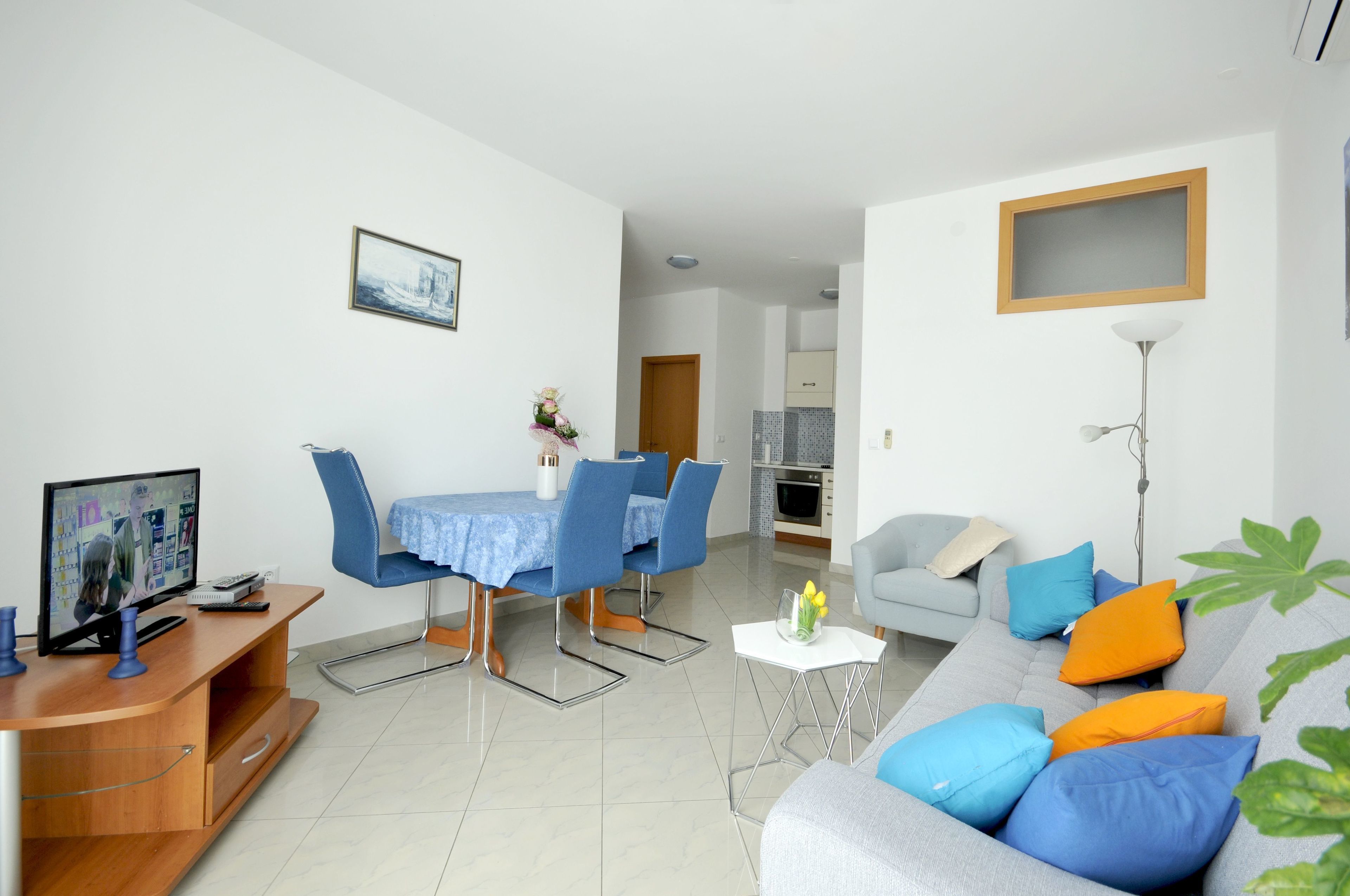 A modern living room with a dining area, a grey sofa with colorful cushions, a television on a wooden stand, and a small table with a vase of flowers.