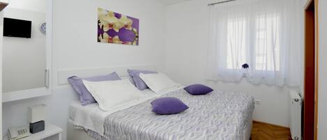 A bedroom with a bed, white walls, a window with white curtains, a mounted air conditioner, a bedside table with a lamp, and a wall clock.