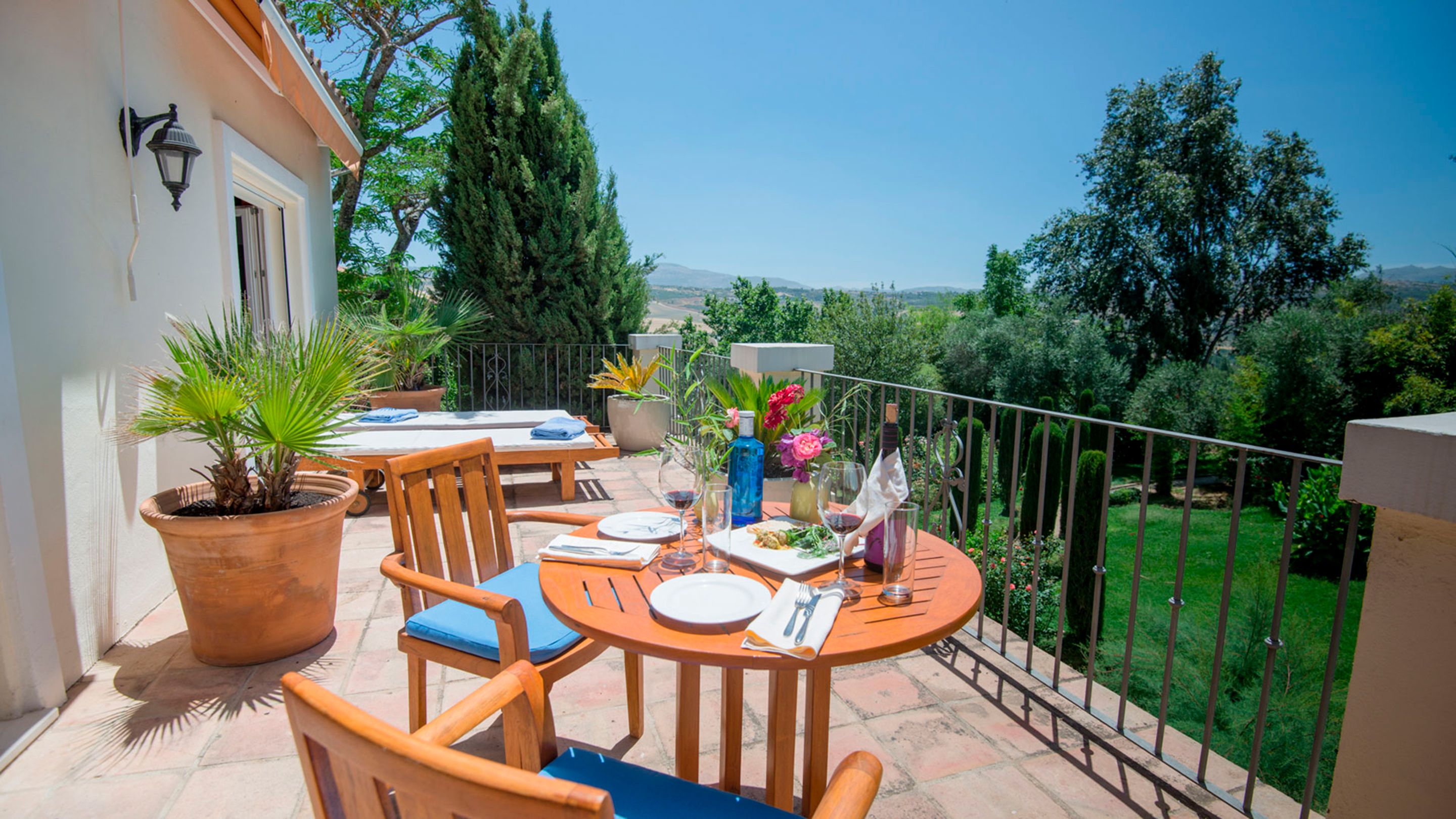 Une terrasse extérieure avec une table dressée pour deux personnes, entourée de plantes en pot et offrant une vue sur la campagne.