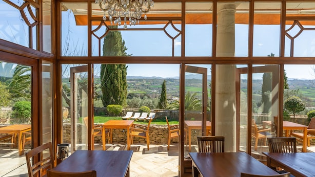 Un vaste espace extérieur aménagé pour les repas, avec des tables et des chaises en bois, de grandes fenêtres offrant une vue sur la campagne et un lustre décoratif.