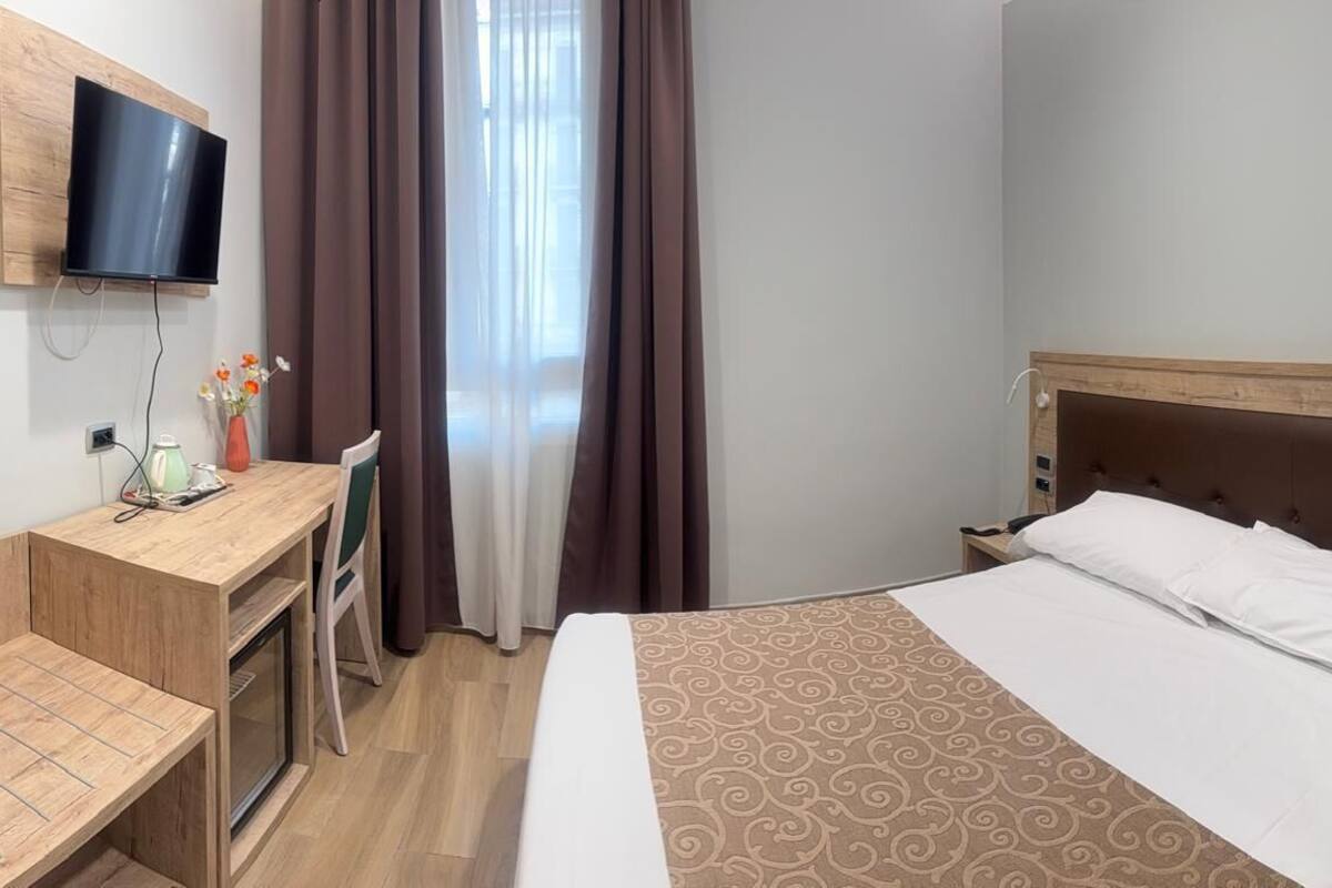 Habitación de hotel con cama, escritorio con televisor de pantalla plana, florero con flores y ventana con cortinas.