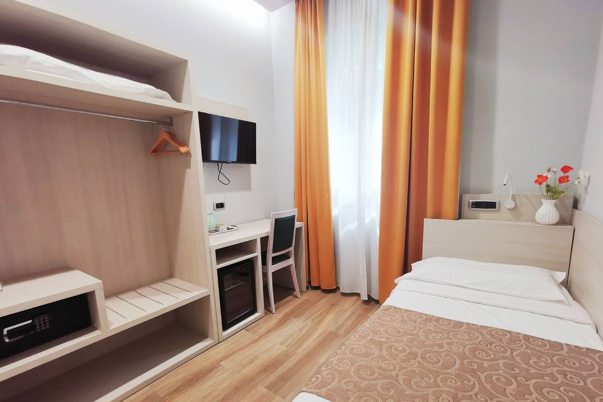 Una habitación de hotel con cama, un escritorio con silla, televisor montado en la pared, armario y ventana con cortinas.