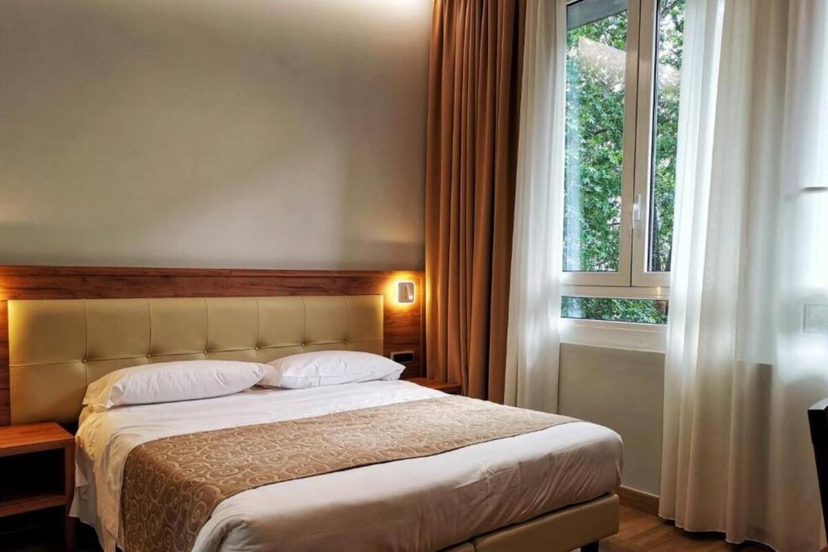 Habitación de hotel con una cama grande, cabecera de madera, dos mesitas de noche y una ventana con cortinas.