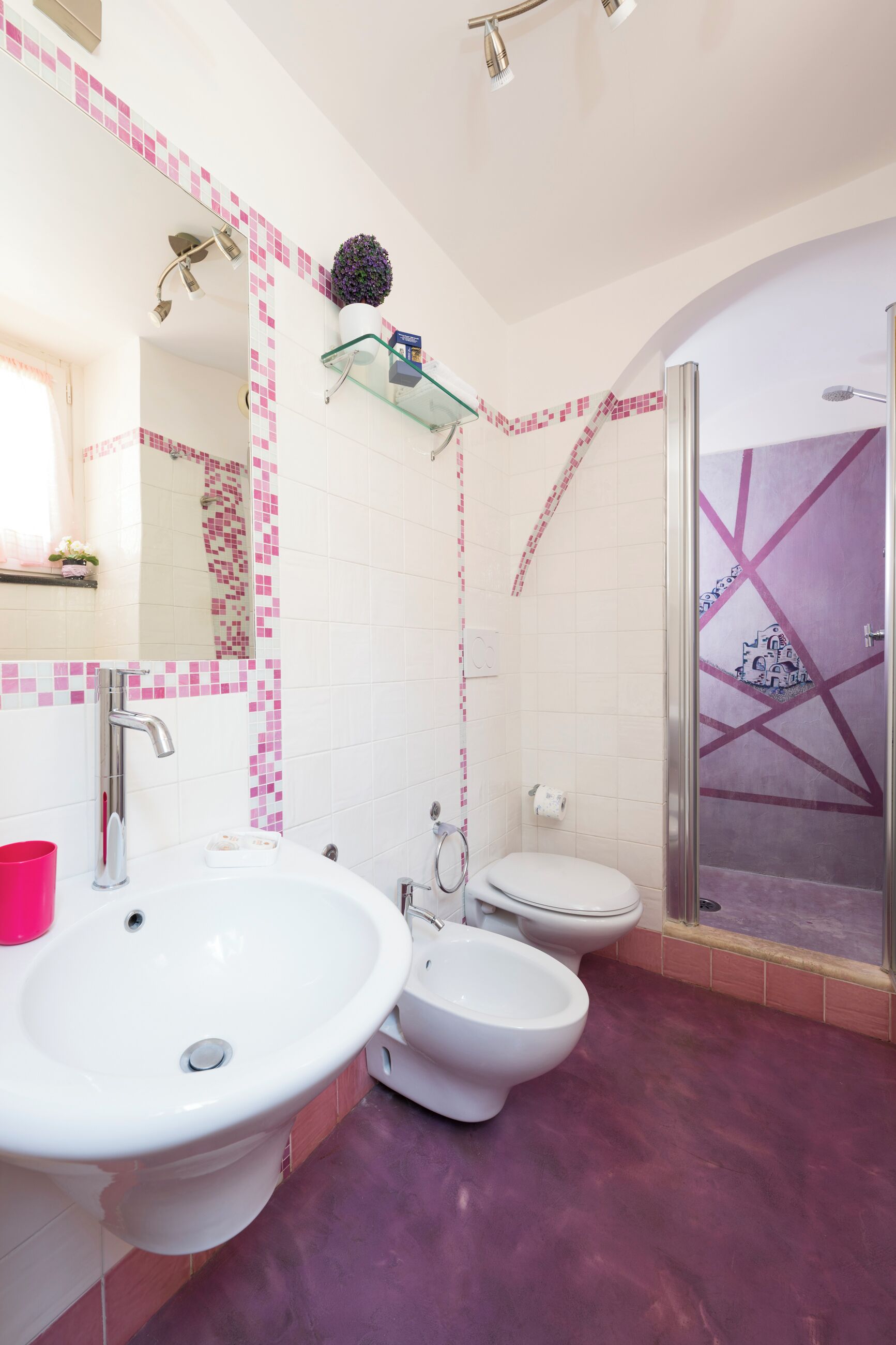 Un baño con un lavamanos blanco, dos inodoros, una ducha y un estante de vidrio con una planta en maceta.
