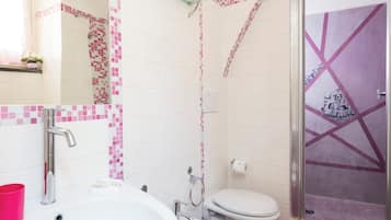 Un baño con un lavamanos blanco, dos inodoros, una ducha y un estante de vidrio con una planta en maceta.