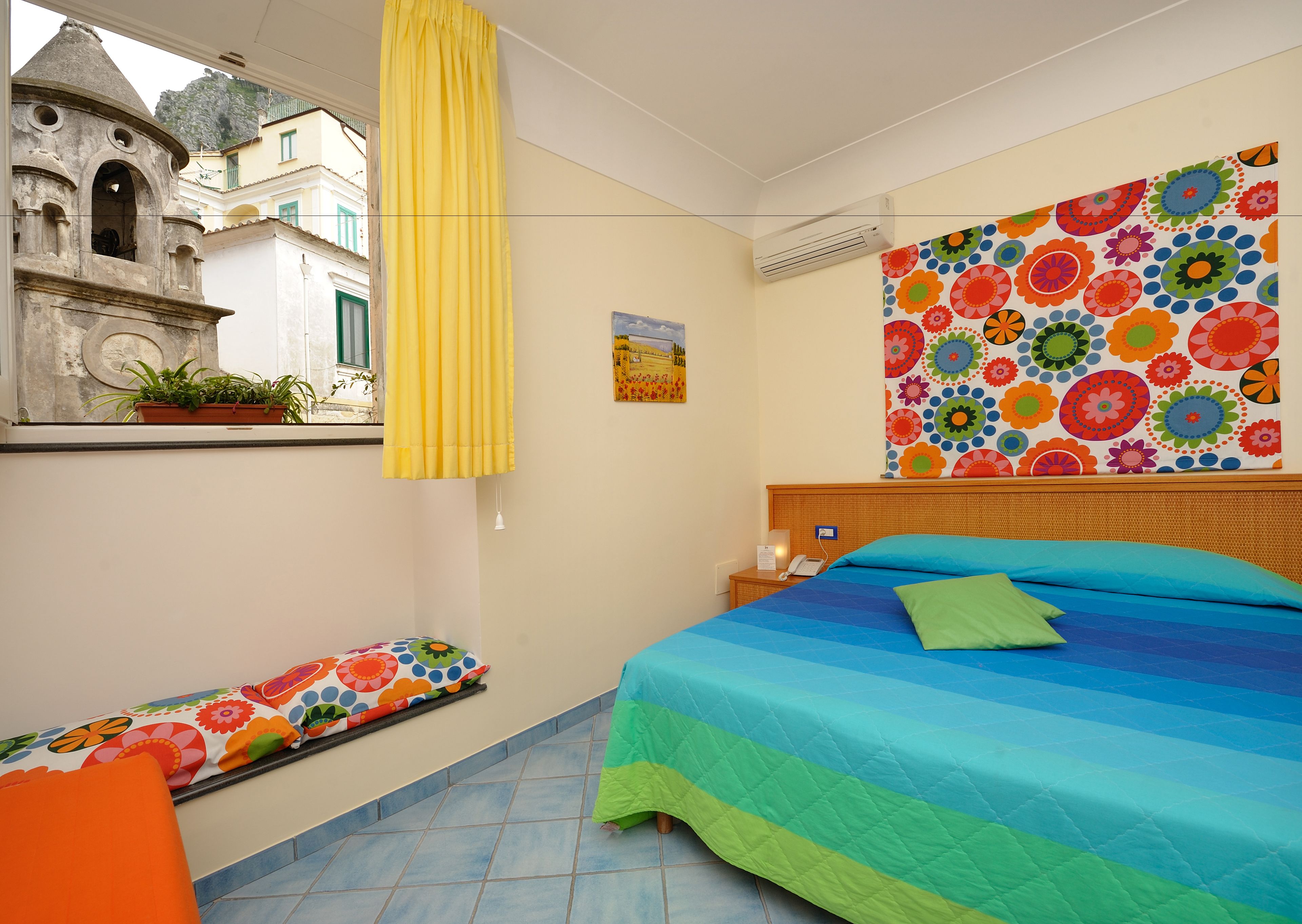 Un dormitorio con cama, una mesita de noche, una ventana con vista a un edificio y un colorido adorno de pared.