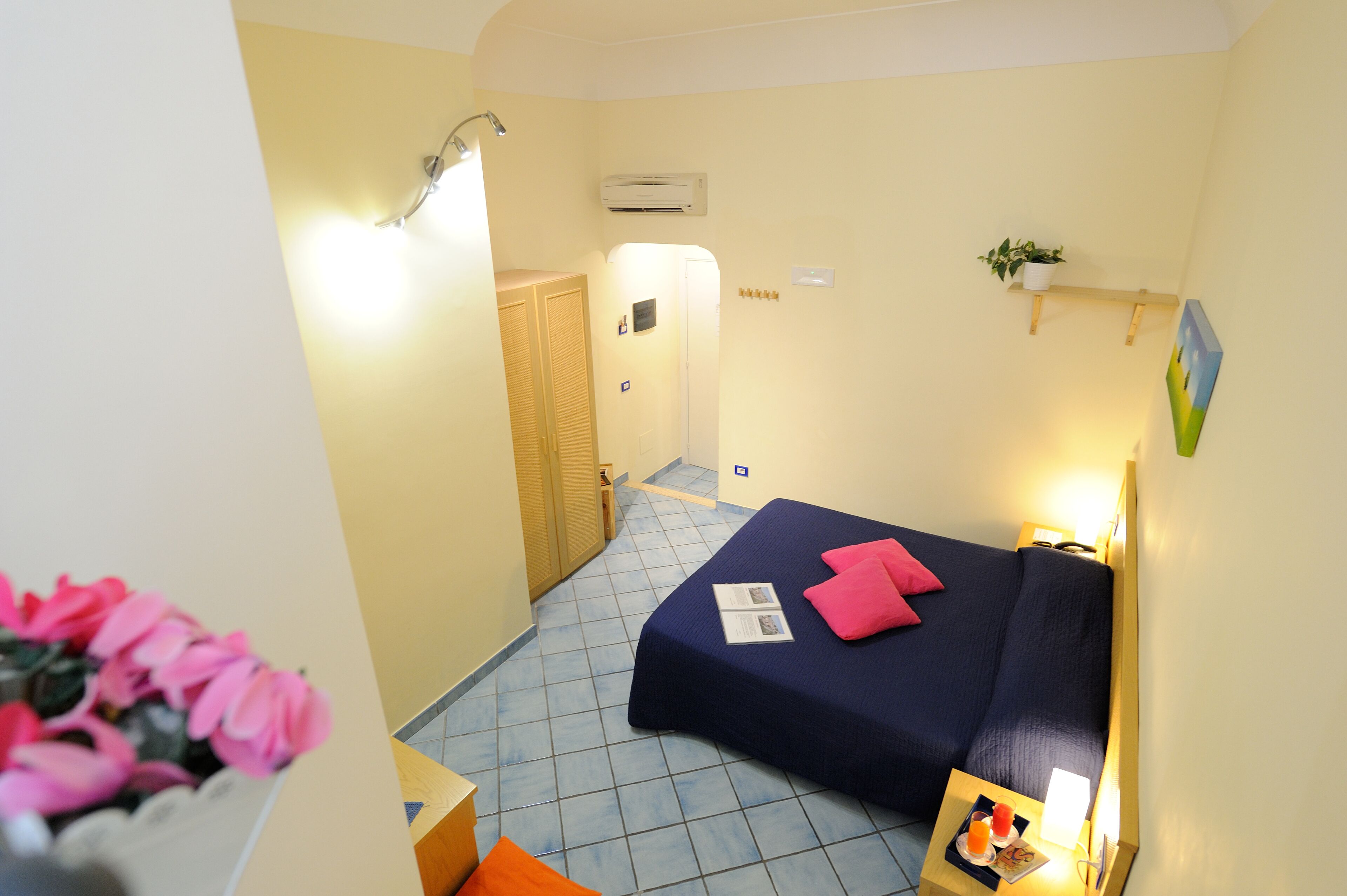 A small, well-lit room with a bed, a bedside table with a lamp, a wall-mounted air conditioner, and a small shelf with a plant.
