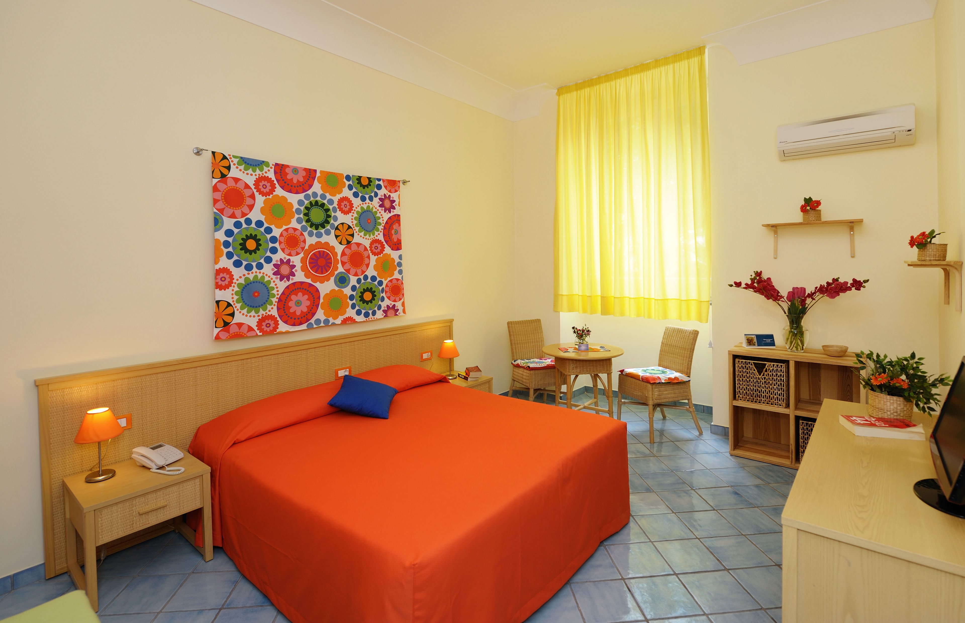 A hotel room with a large bed, a desk, a chair, a small table, a colorful artwork on the wall, and a window with yellow curtains.