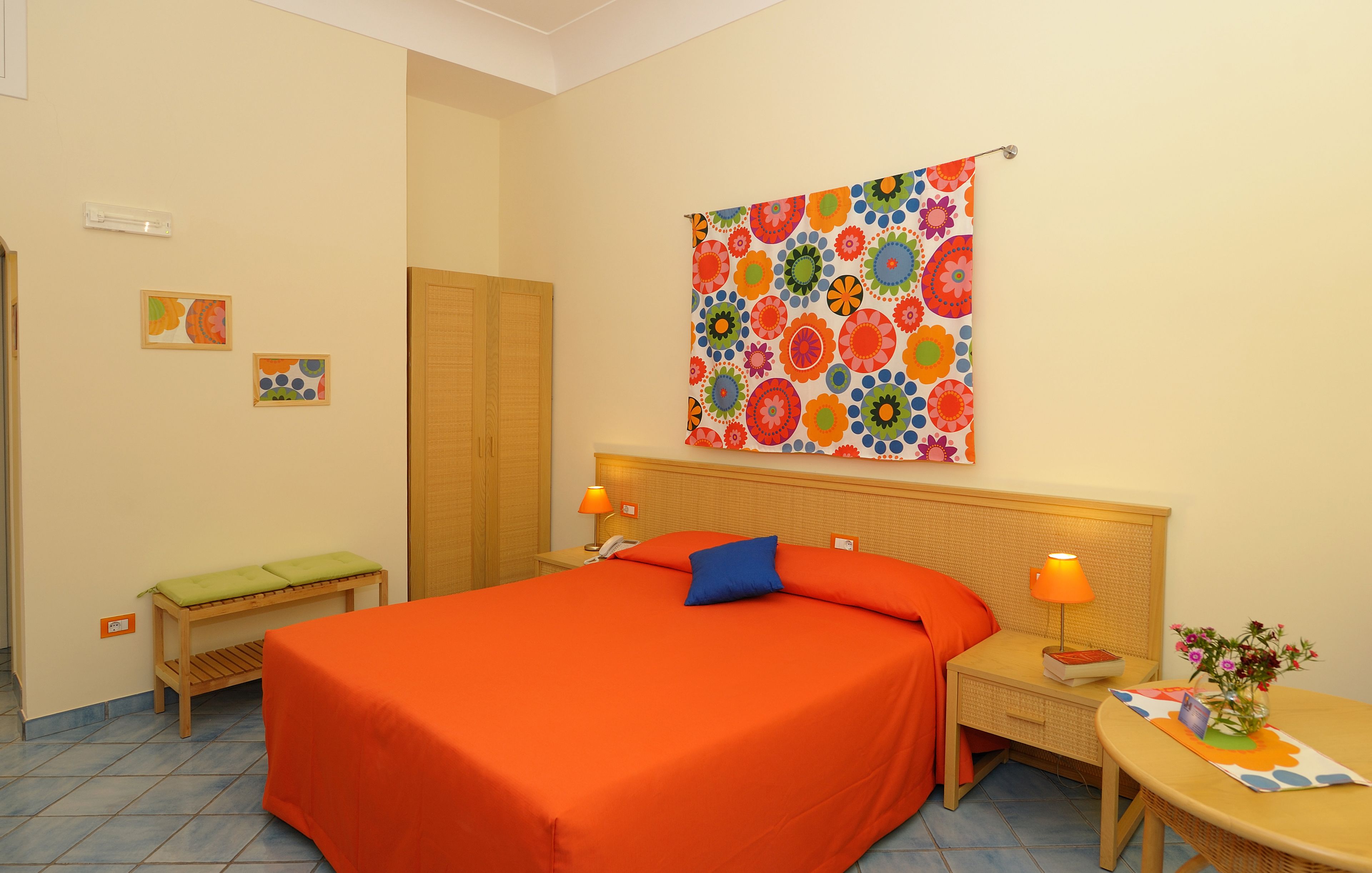 A hotel room with a large bed, a nightstand, a small table, and a colorful floral tapestry on the wall.