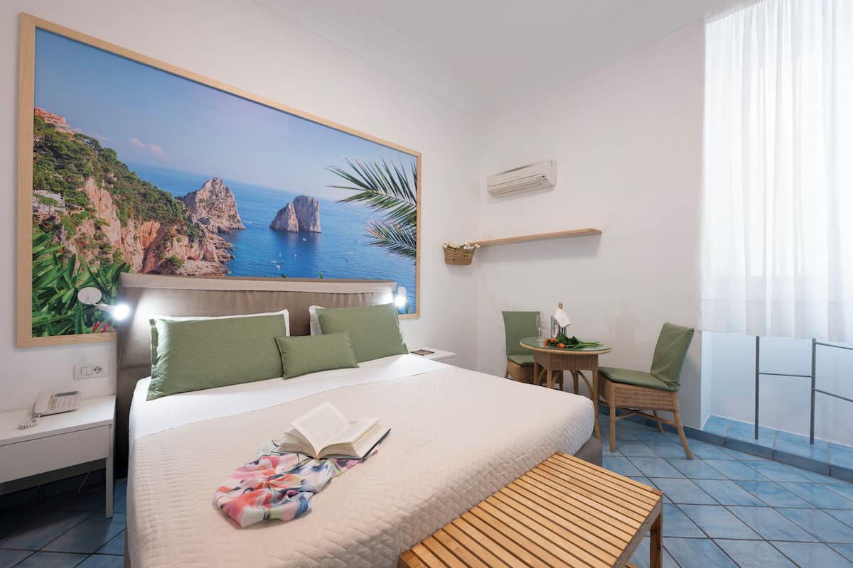 A hotel room with a bed, a desk, a chair, a small table, and a large painting of a coastal landscape.