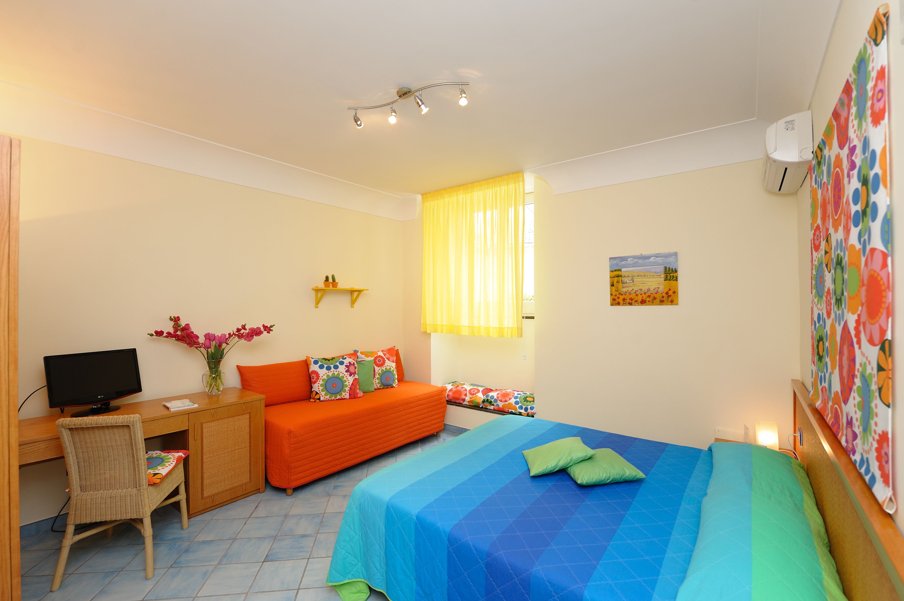 Une chambre à coucher avec un lit, un bureau avec un ordinateur, une chaise, un canapé et une fenêtre avec des rideaux.
