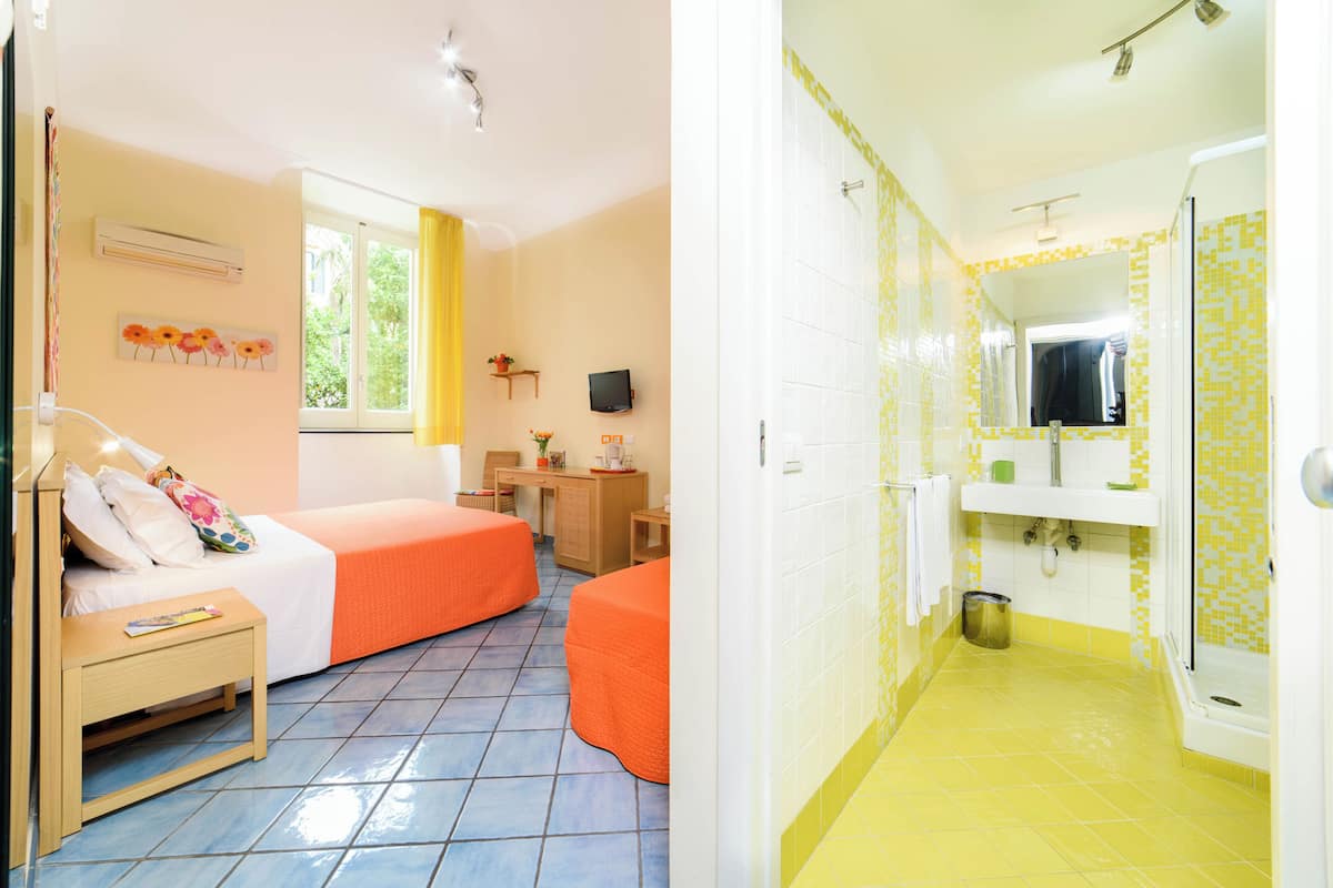 A room with two beds, a desk, a TV, and a window with yellow curtains.