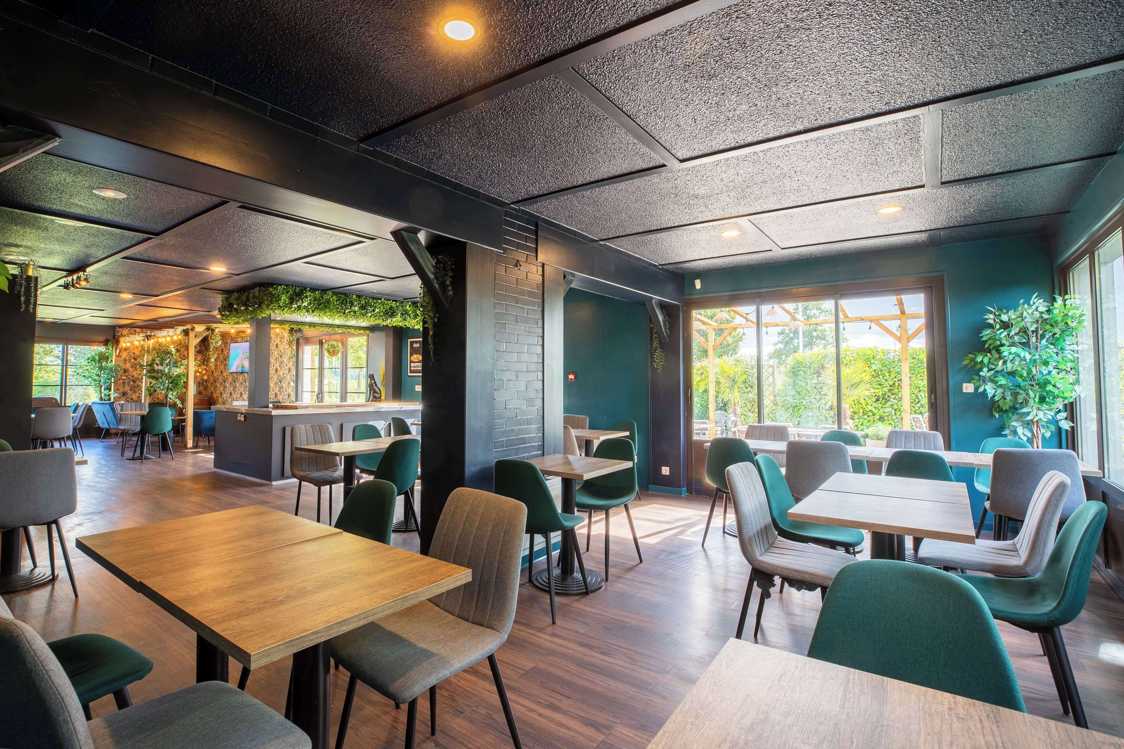 Un restaurante moderno con mesas de madera, sillas verdes, ventanas amplias y una pared de ladrillo con plantas.