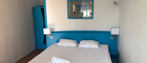 A hotel room with a large bed, two pillows, a blue headboard, a mirror, and a wall-mounted light.