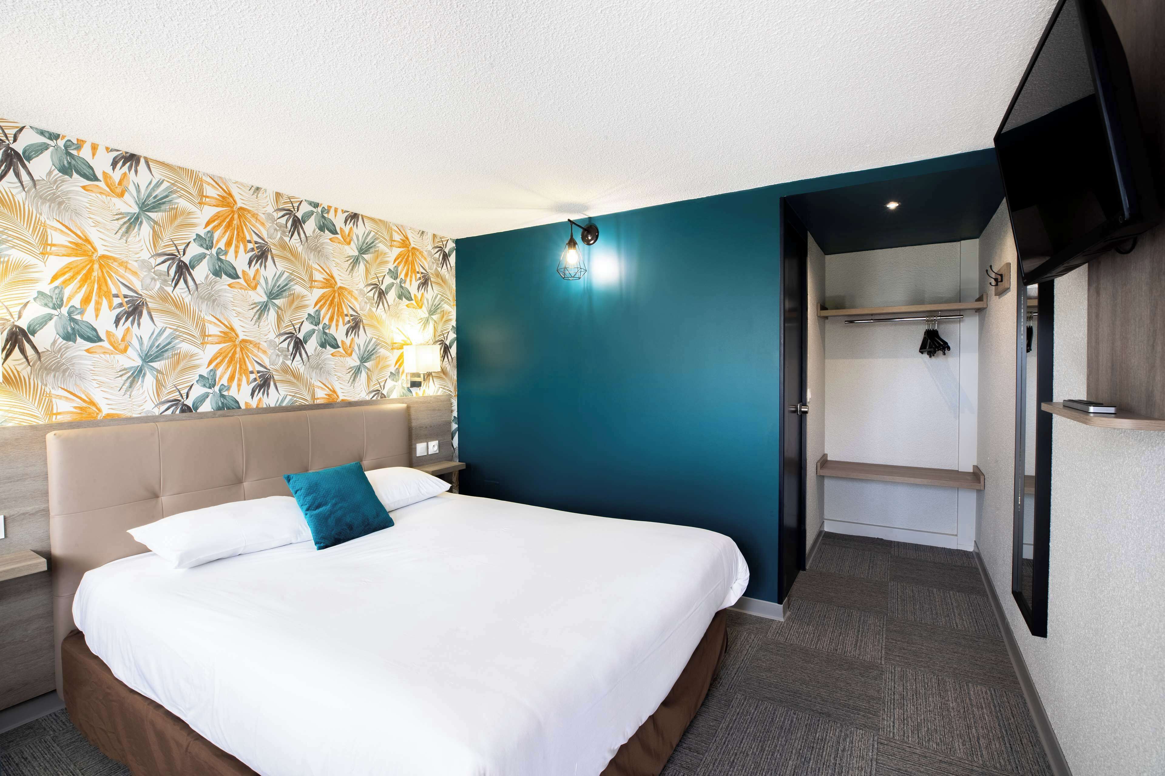 Une chambre d’hôtel comprenant un lit, une table de chevet, un mur orné d’un motif de feuilles tropicales, un mur d’accent de couleur turquoise, un placard et un téléviseur fixé au mur.