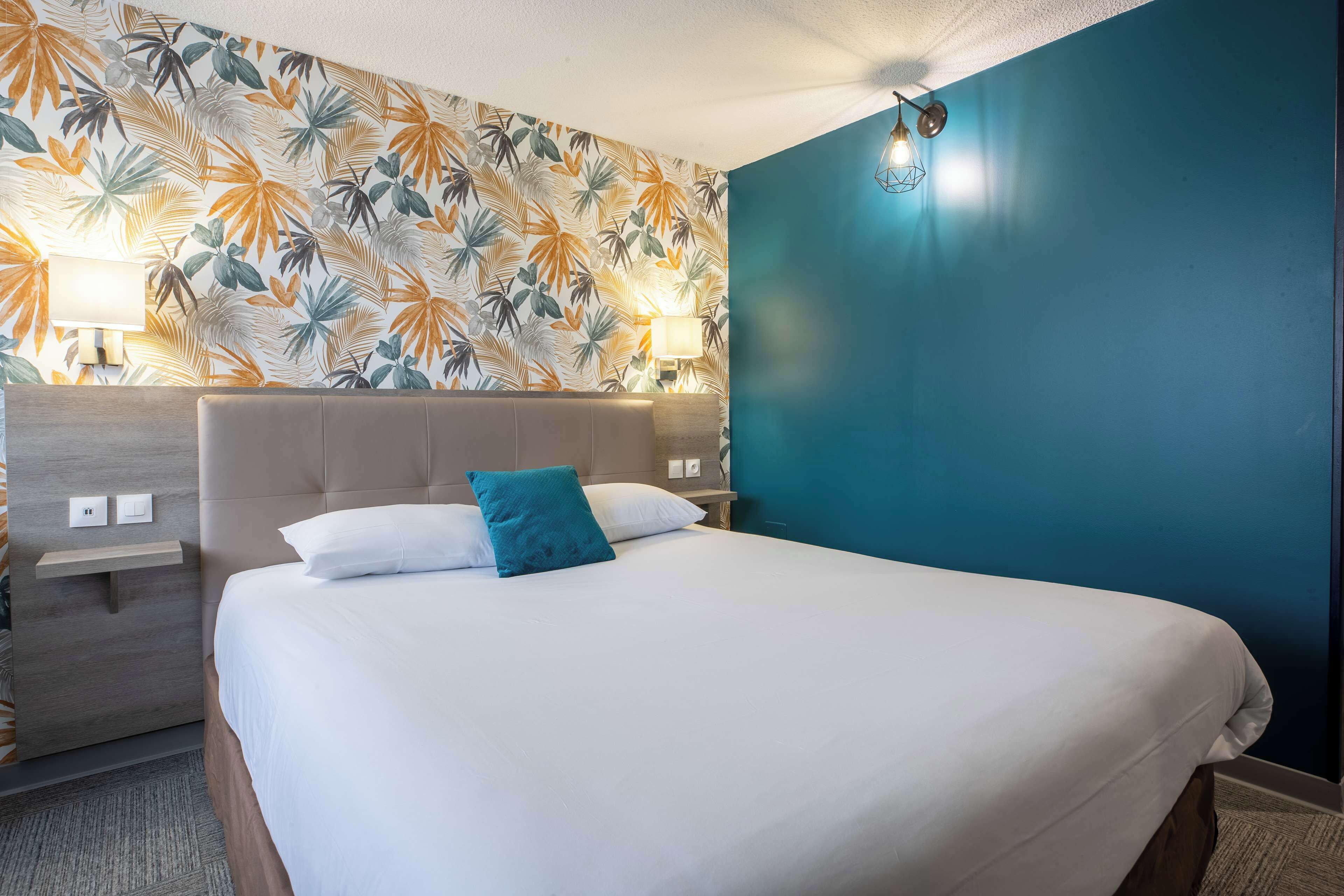 A hotel room with a bed, a nightstand, a lamp, and a wall with a tropical leaf pattern.