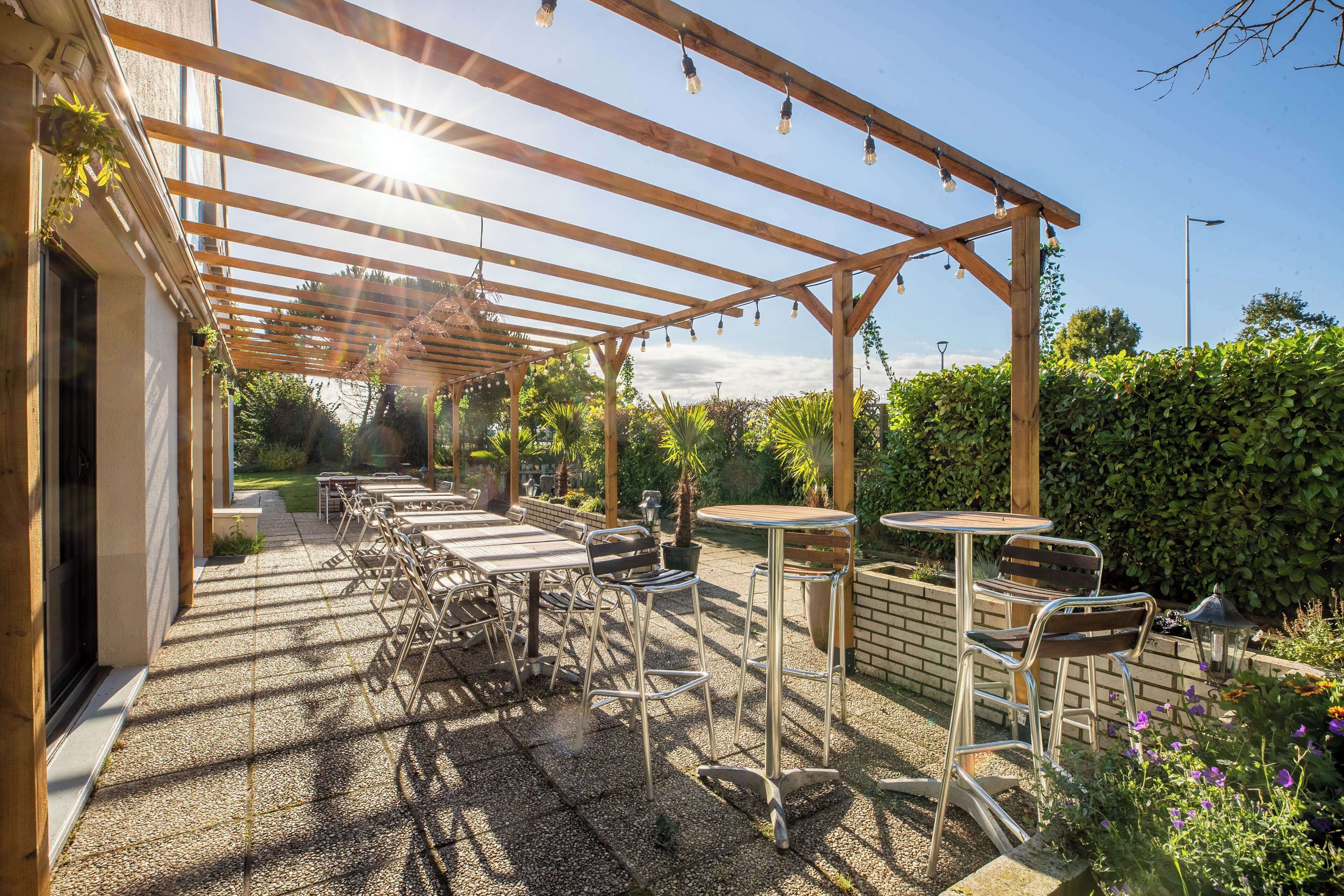 A covered outdoor patio with tables and chairs, a pergola with string lights, and a garden area with plants and a hedge.
