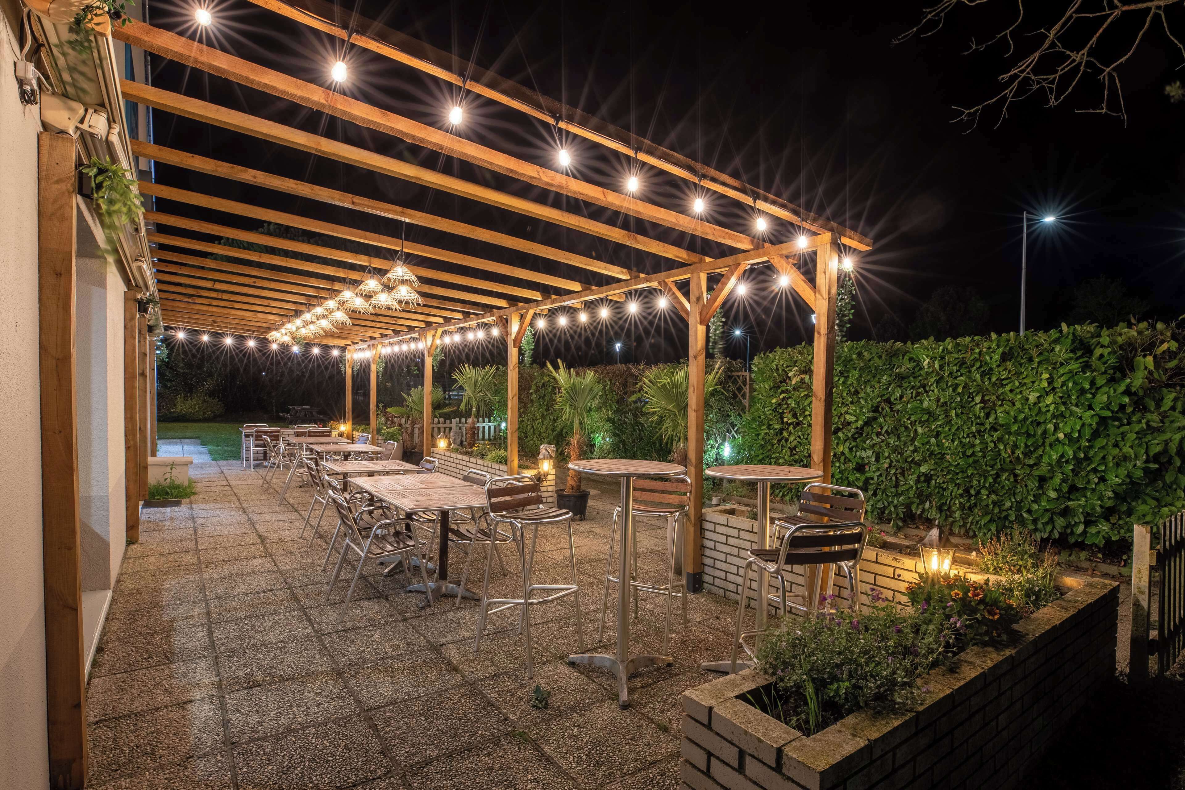Un patio exterior con pérgola de madera, luces colgantes y mesas y sillas.