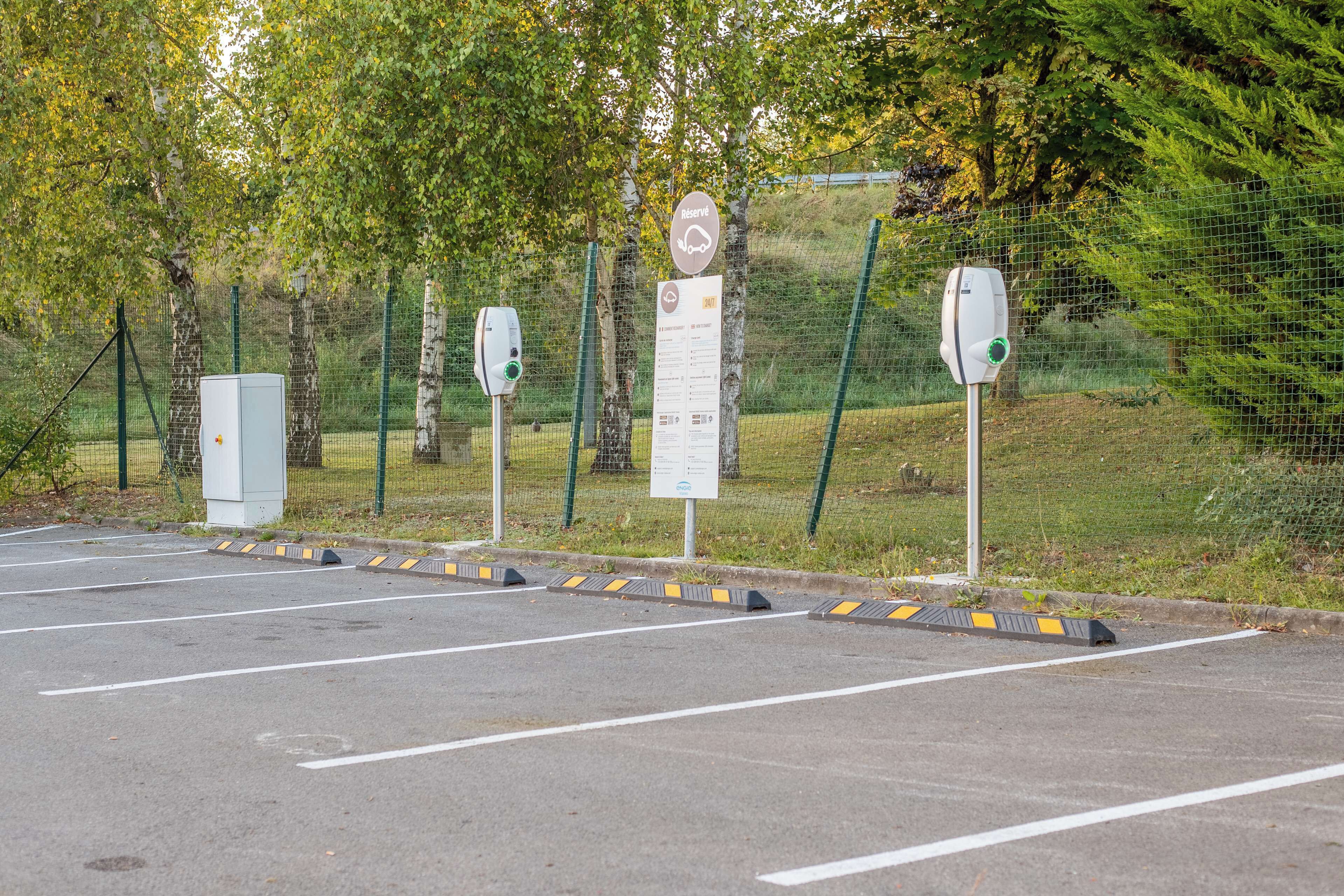 Un estacionamiento con estaciones de carga para vehículos eléctricos, un cartel y un área cercada con árboles.