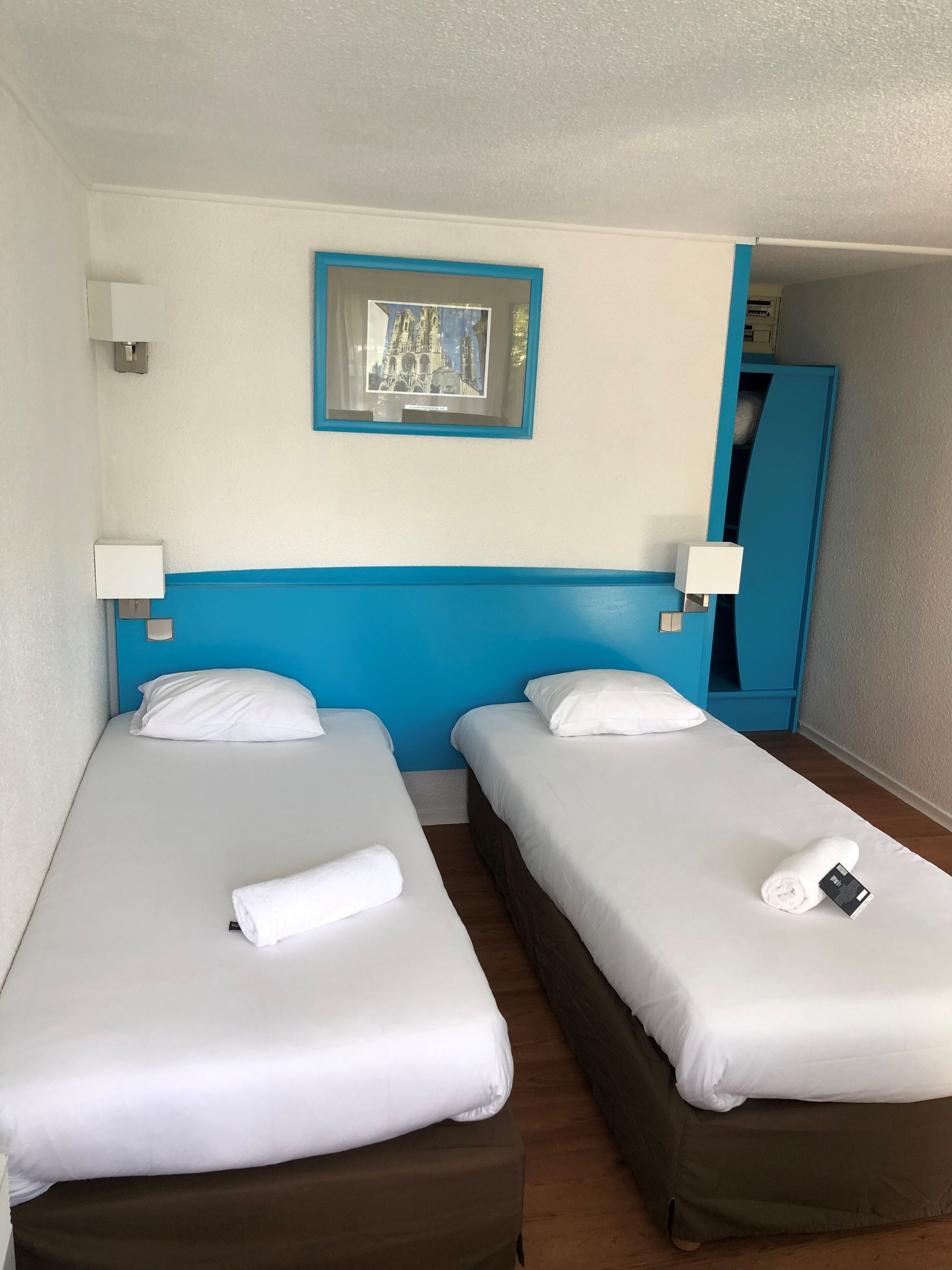 Habitación de hotel con dos camas individuales, ropa de cama blanca, cabecera azul, un cuadro en la pared y un armario azul.