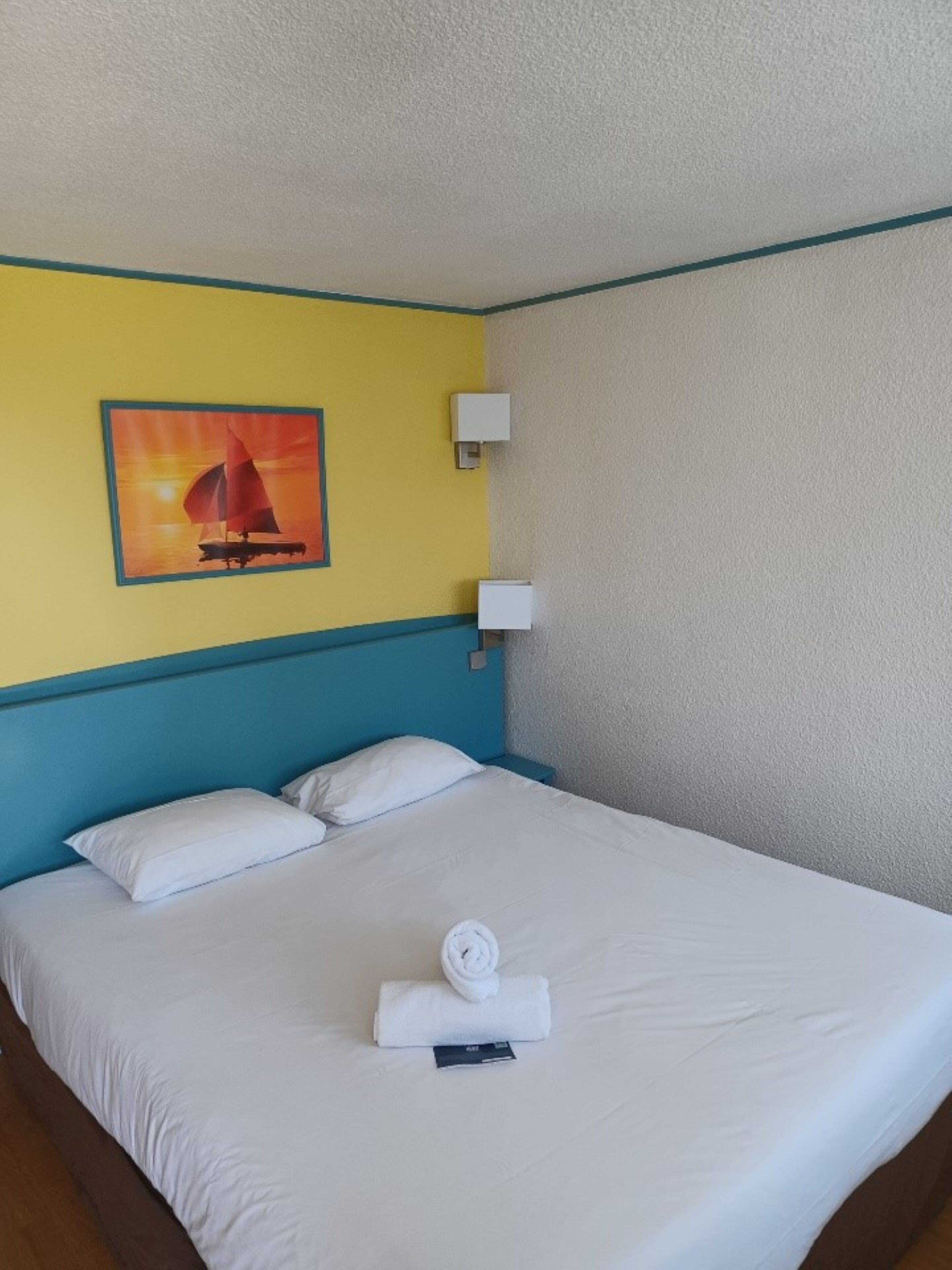 A hotel room with a bed, white linens, a painting of a sailboat, and a wall-mounted light.
