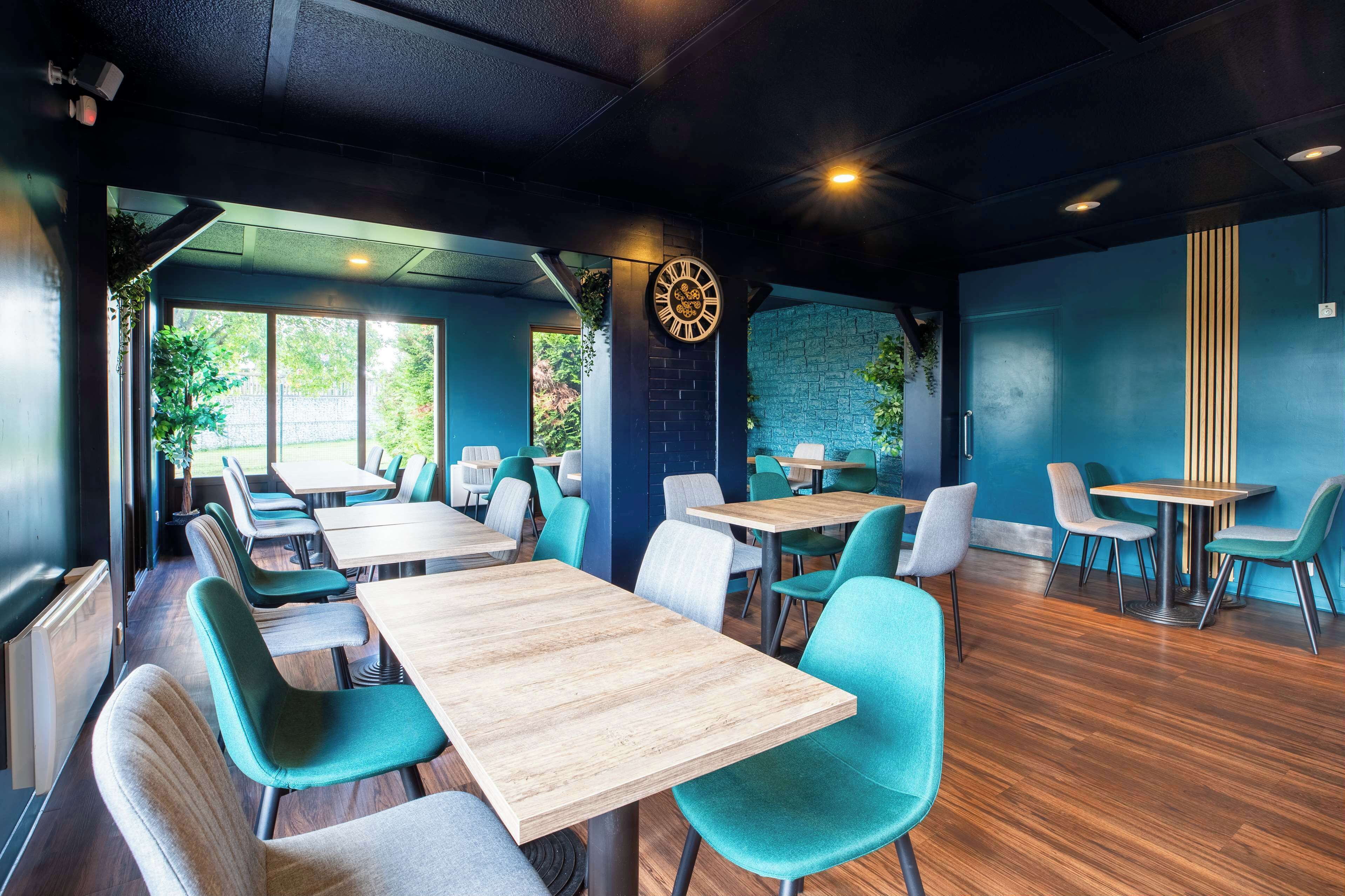 Un interior de restaurante moderno con mesas de madera y sillas color turquesa, un reloj grande en la pared y una pared de acento de ladrillo.