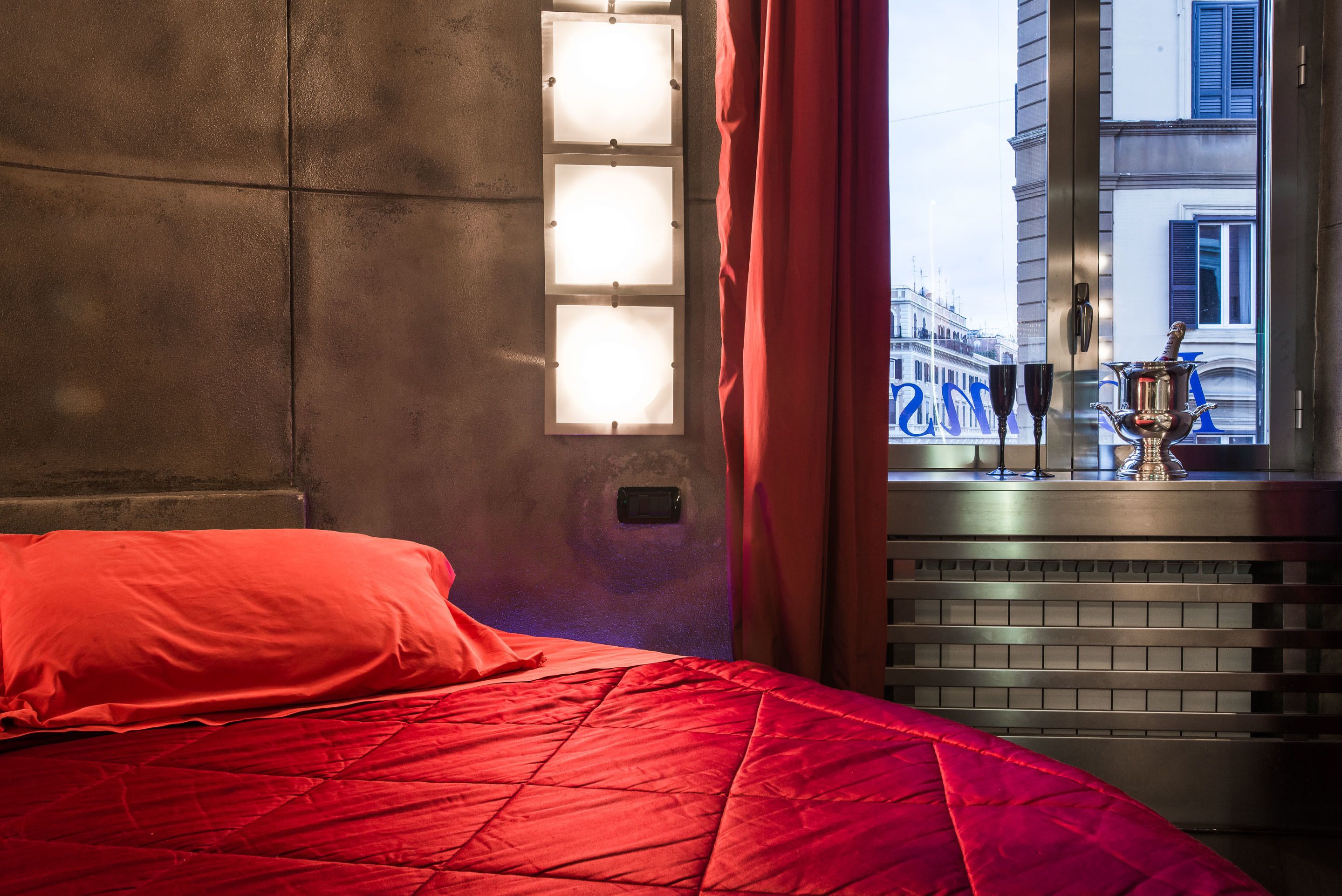 Une chambre d’hôtel avec une couvre-lit rouge, une fenêtre avec des rideaux et une vue sur un bâtiment à l’extérieur.