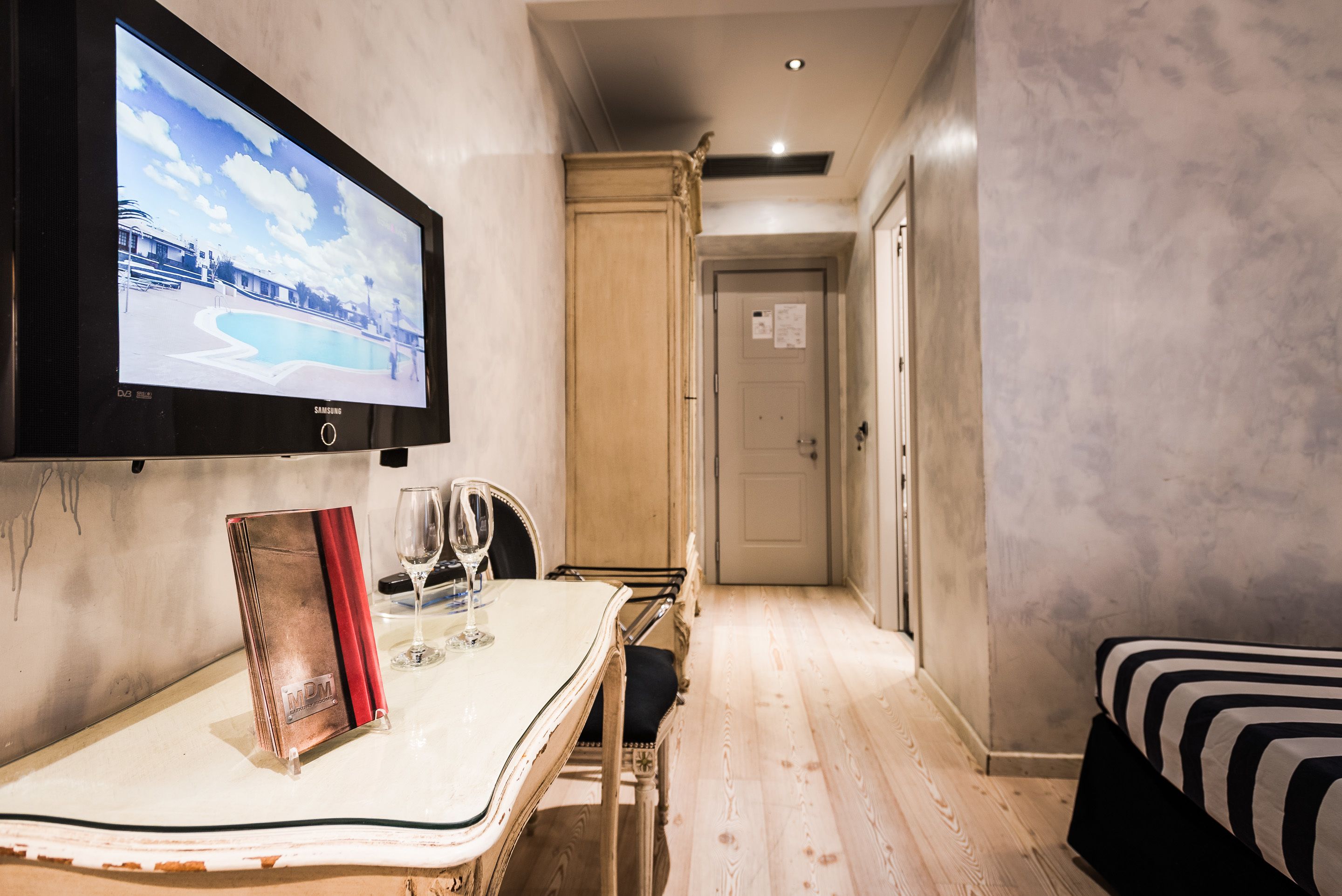 Habitación de hotel con televisor montado en la pared, una mesa de comedor con un menú y una copa de vino, una cama con ropa de cama a rayas y piso de madera.