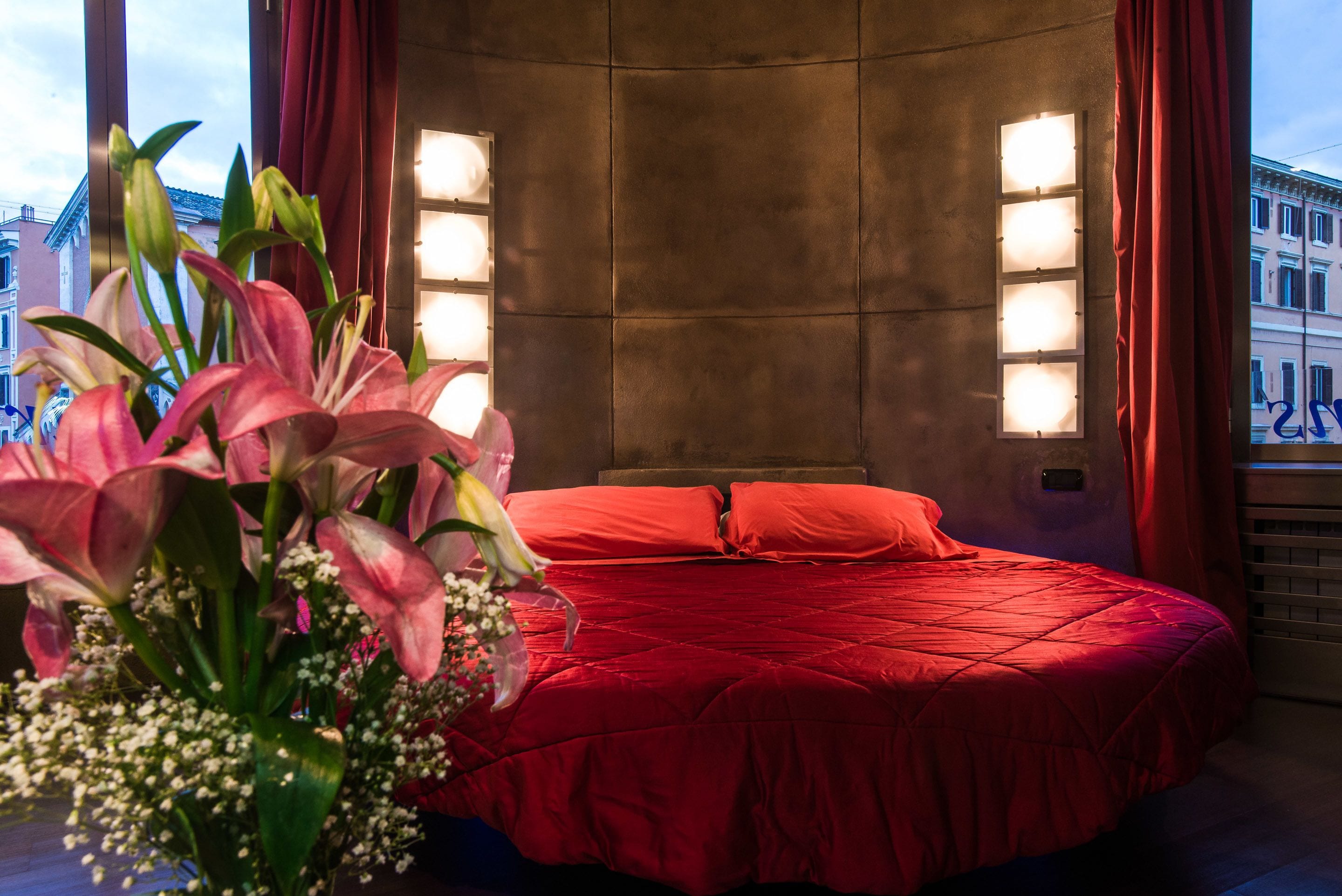 Camera da letto con copripiumino rosso, un bouquet di fiori e vista sulla città dalla finestra.