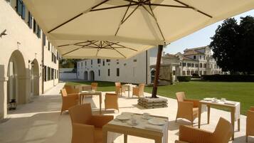 An outdoor patio with wicker furniture and umbrellas, a white building in the background, and a well-maintained lawn.