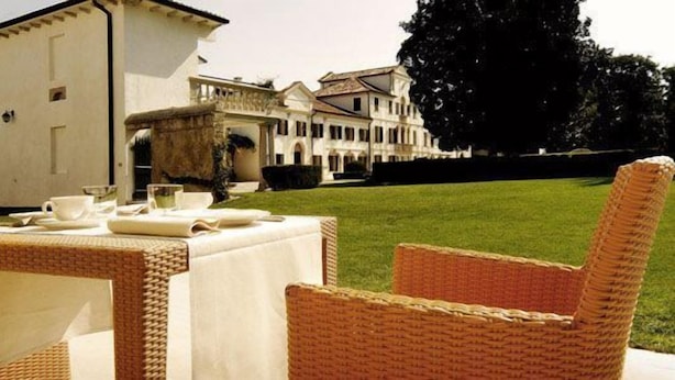 A dining area with wicker chairs and a table set for a meal, overlooking a large building with a well-maintained lawn. - Villa Toderini Agriturismo (Codogne)