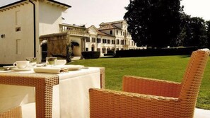 A dining area with wicker chairs and a table set for a meal, overlooking a large building with a well-maintained lawn. - Villa Toderini Agriturismo (Codogne)