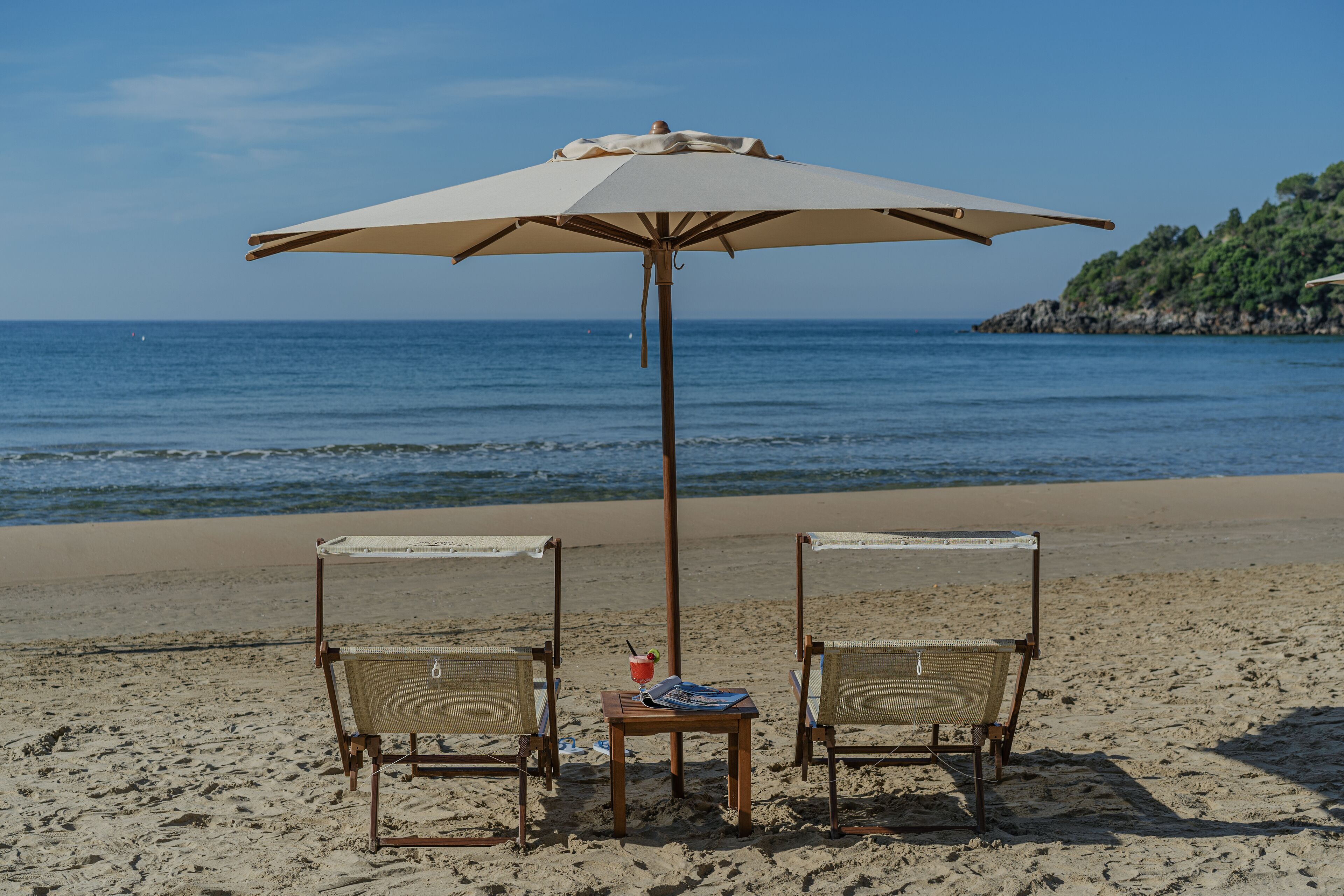 Una spiaggia con due sedie in vimini sotto un ombrellone grande, un tavolino con una bevanda e l'oceano sullo sfondo.