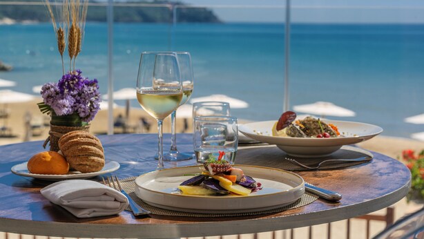 Una mesa con comida, una copa de vino blanco y vista al mar.