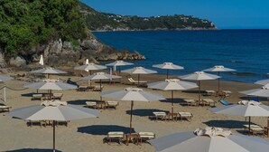 Una playa con hileras de sombrillas y tumbonas, un litoral rocoso y vistas al mar y una colina distante con edificios.